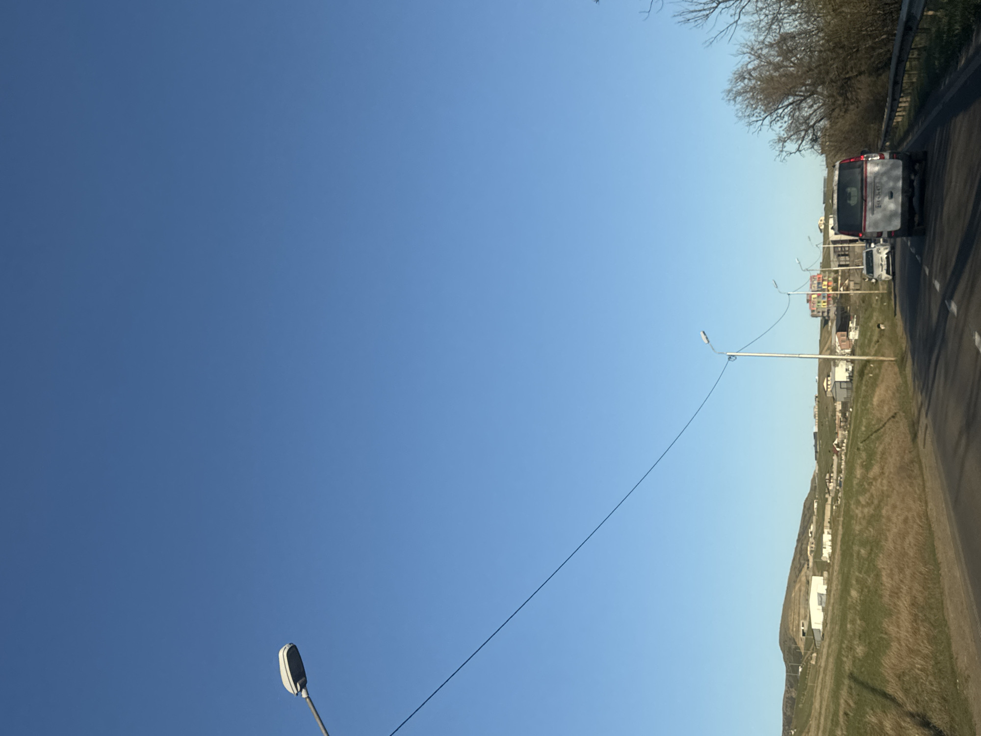 The image depicts a road with a white van traveling along it. Streetlights line the road, with a power line visible in the sky. Buildings are scattered on a grassy hill adjacent to the road. The sky is clear blue. The location is in the city of Tbilisi, Georgia.