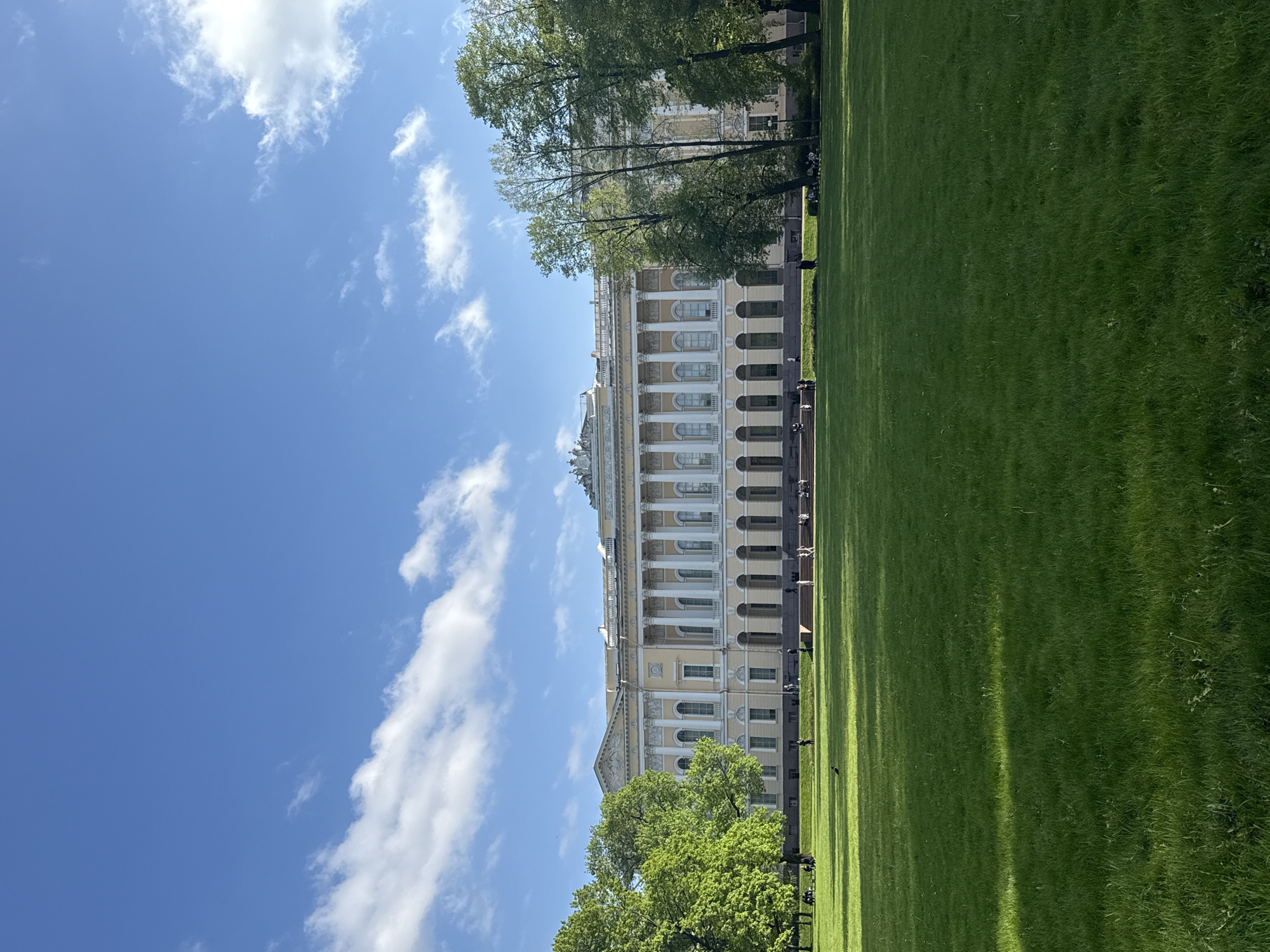 The image shows the facade of the Russian Museum in Saint Petersburg, Russia, viewed from a park. The building is light yellow with prominent white columns. A green lawn stretches in front of the museum, and the sky above is blue with scattered white clouds. Trees partially obscure the lower portion of the building. A few people are visible in the park, seated or walking. The lighting suggests it is daytime with bright sunlight.