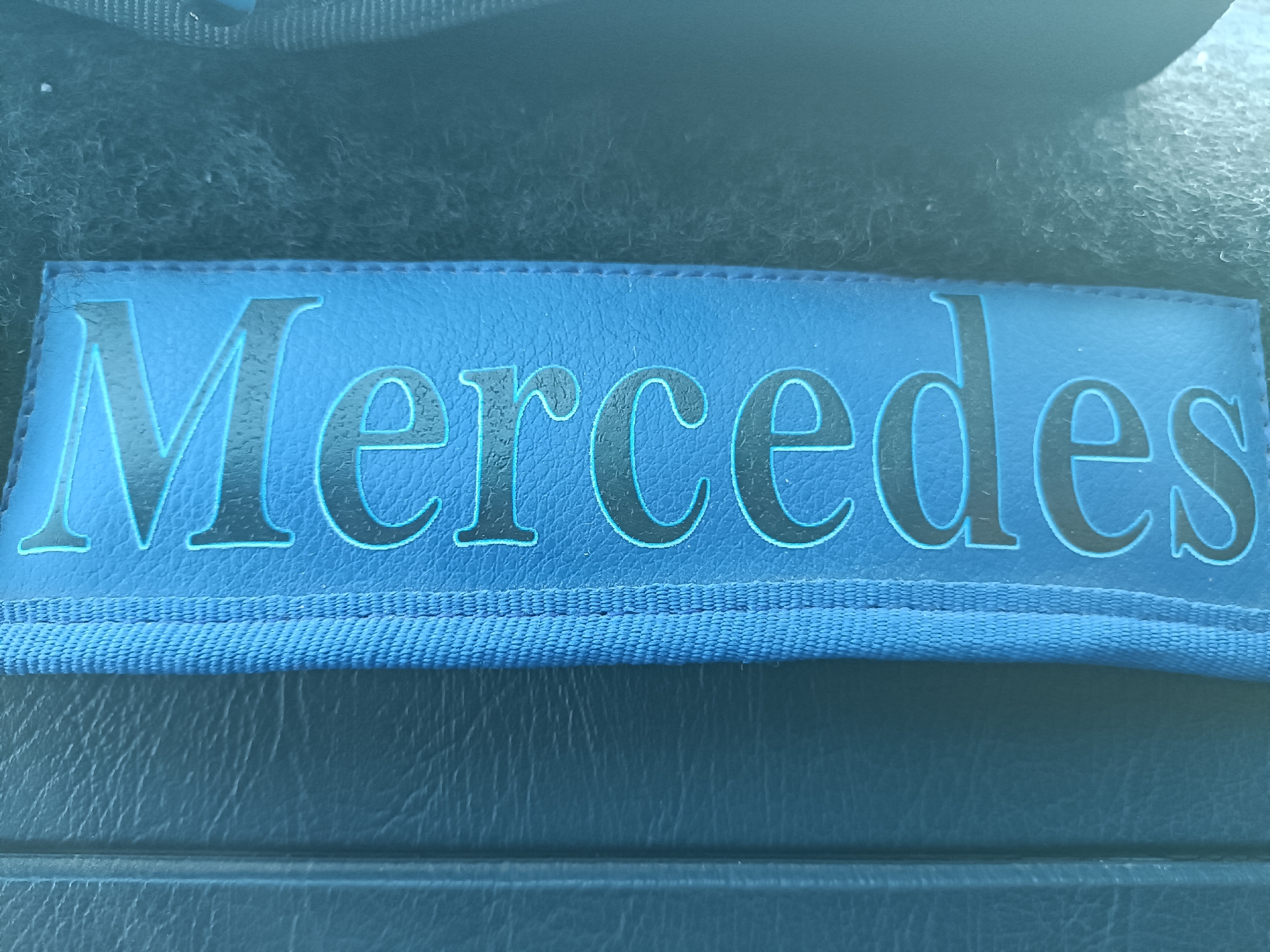 The image shows a rectangular blue patch with the word "Mercedes" written in black letters with a light blue outline. The patch appears to be made of leather or a similar material, and it is lying on a dark surface with a textured pattern. The top portion of the image features a fuzzy or fabric-like material, and the lighting is somewhat dim.