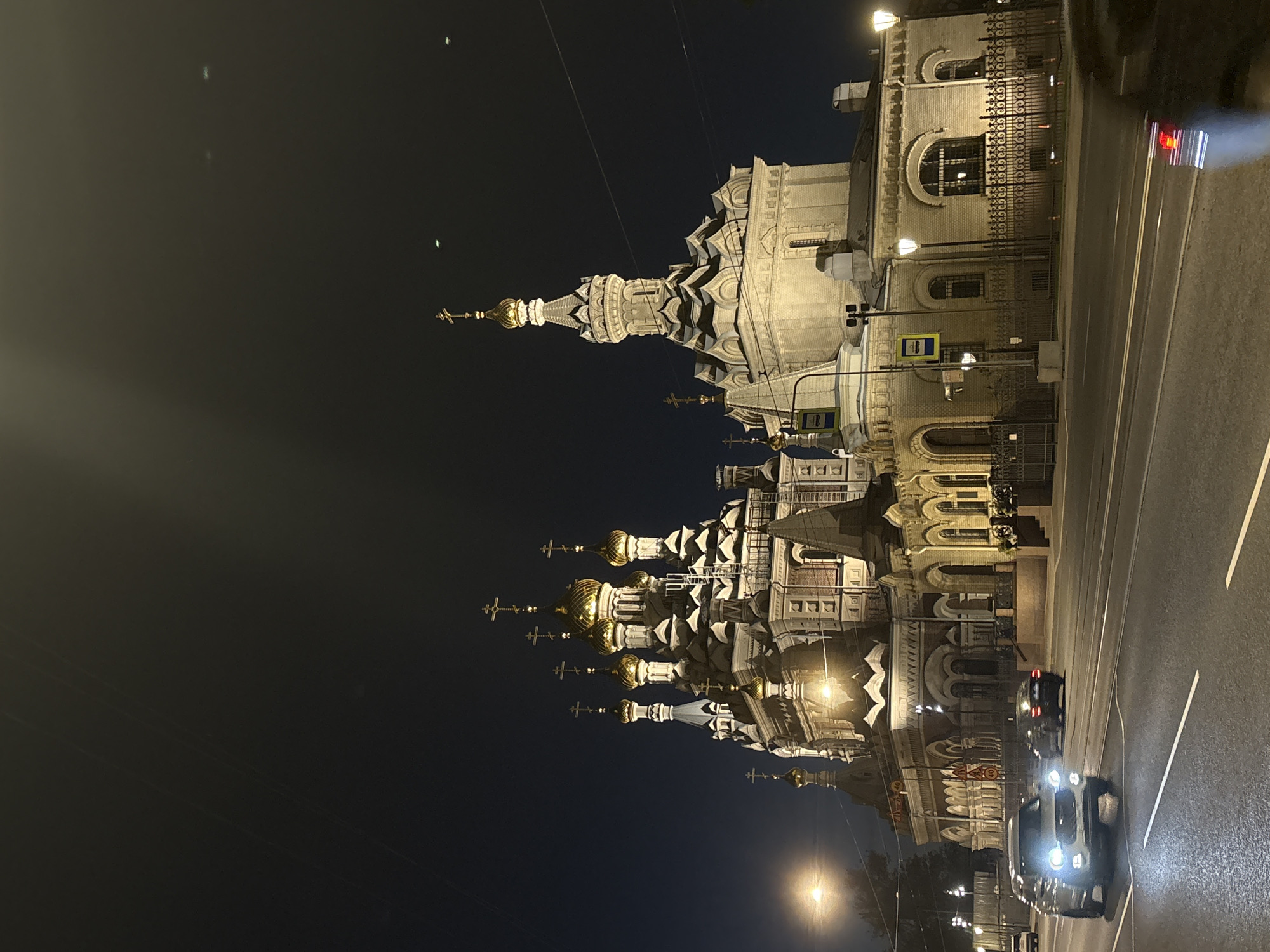 This is a nighttime, eye-level, exterior shot of the Chesme Church, officially the Church of St. John the Forerunner at Cherepovo, in Saint Petersburg, Russia. The church is illuminated and its red brick and white stucco facade, along with its numerous golden domes and crosses, are clearly visible against the dark night sky. In the foreground, a multi-lane road is visible, with several cars driving on it. The closest car is a silver SUV with its headlights on. The church is located on the right side of the road from the perspective of the viewer. The scene appears to be a street in a city, with streetlights and overhead wires also visible. The overall mood of the image is serene and majestic, highlighting the beauty of the church at night.
