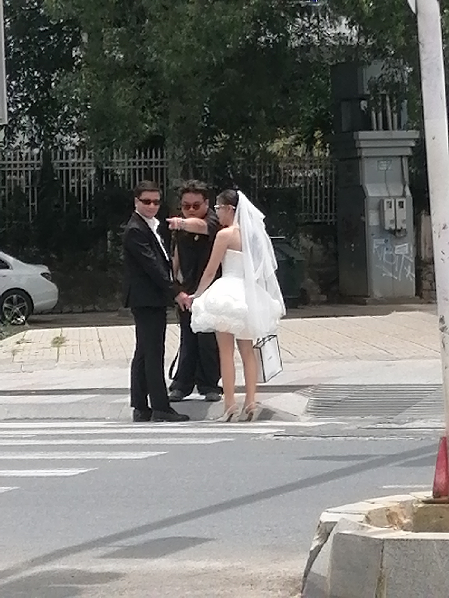 In Da Lat, Vietnam, at a pedestrian crossing, three individuals stand near a white vehicle. A man in a black suit and sunglasses, with his hands clasped, faces the viewer. Beside him, another man, also wearing sunglasses, points with his left hand towards something out of frame. A woman, dressed in a short white wedding dress and veil, stands to the right, holding a white shopping bag. The environment includes trees, a fence, and a utility box.