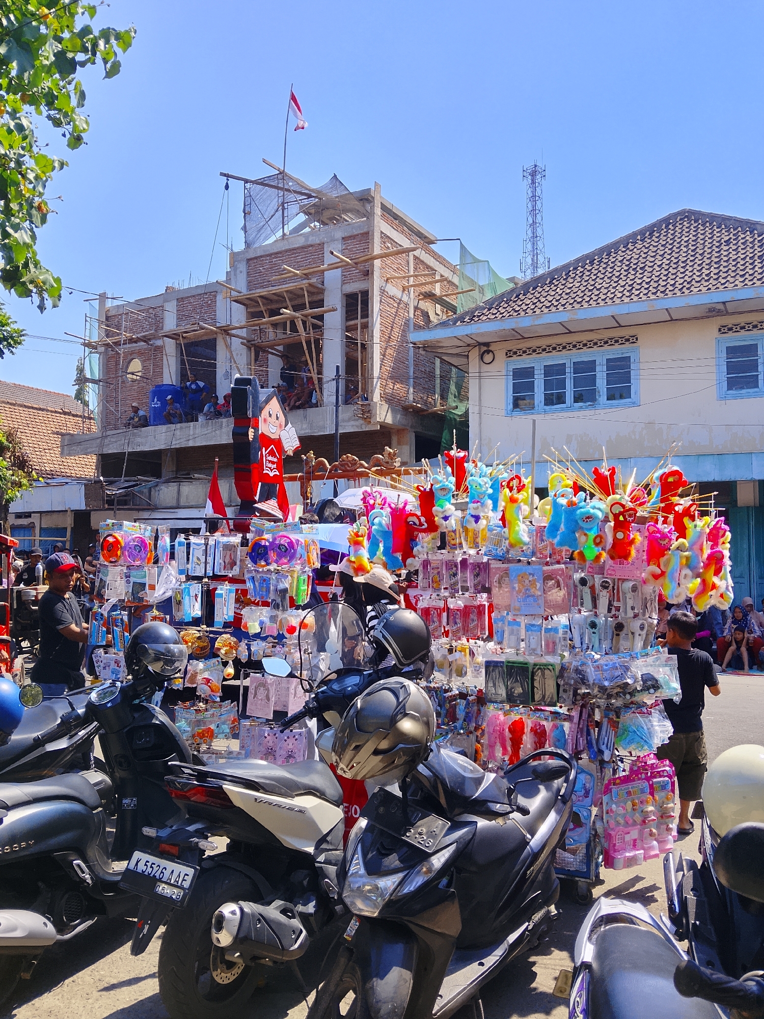 In the city of Ngelo, Indonesia, on a sunny day, a street vendor displays a variety of colorful toys, including balloons and plush figures, in front of a building under construction. Several motorcycles are parked nearby, one with a license plate that reads "K 5526 AAE". People are seen gathered around the stall and in the background, some seated and others standing. The scene is characterized by bright daylight and a clear blue sky, with a prominent cell tower visible in the distance.