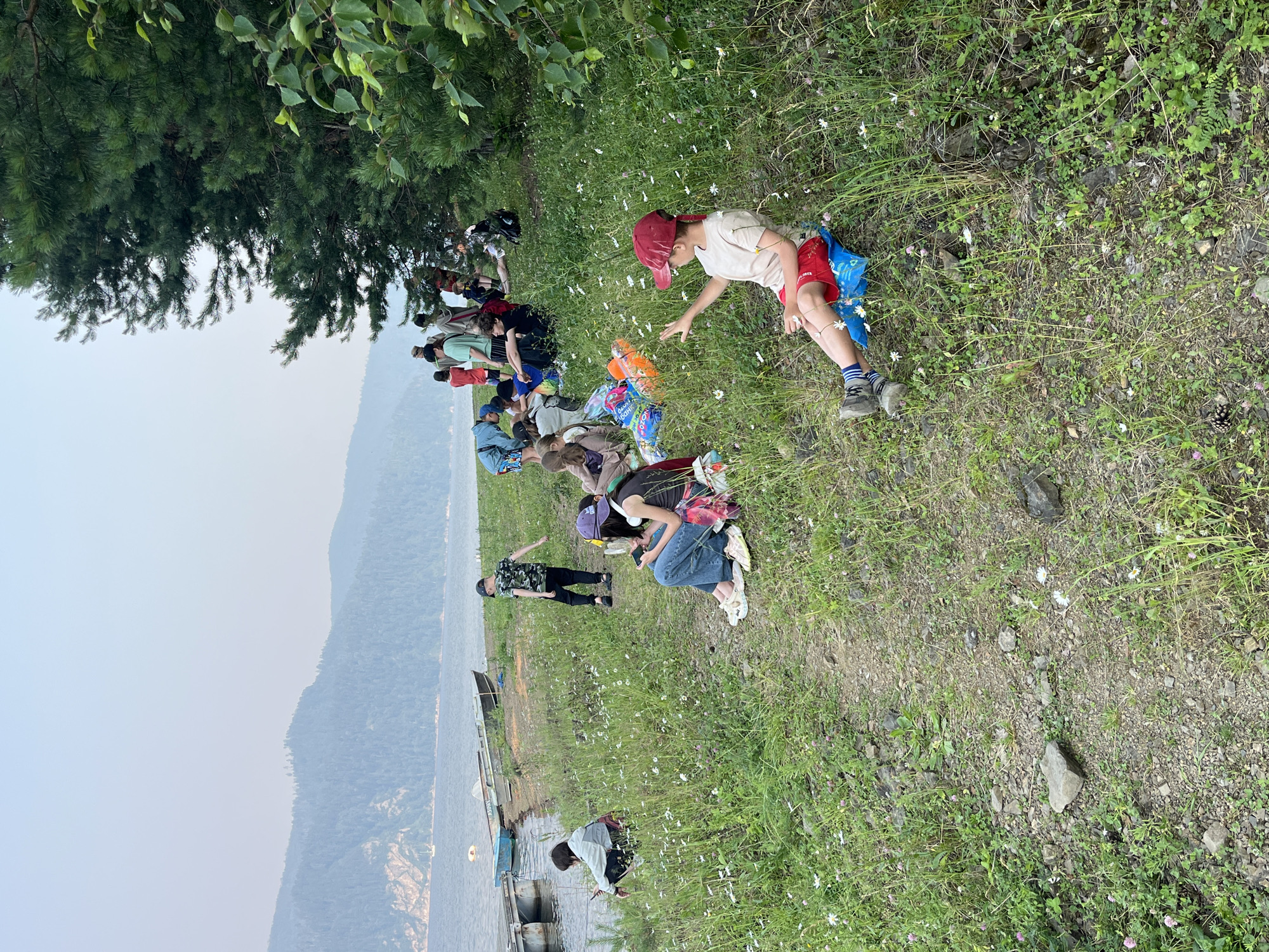 The image depicts a group of people, predominantly children, gathered near a body of water. The setting appears to be a natural, grassy area with a path leading down to the water's edge. Mountains covered in trees are visible in the background across the water, and a hazy sky suggests either early morning or late evening light, or possibly atmospheric conditions like smoke. Several boats are moored at a dock on the left side of the image.
In the foreground, a young boy wearing a red cap and red shorts sits on a blue cushion, gesturing with his right hand. To his left, a person with a purple hat and headphones sits on the ground, looking at a phone. Further left, a person in a light-colored jacket is crouched near the water, also looking at a phone. In the middle ground, a child in a camouflage shirt walks away from the camera. Other individuals and groups of people are scattered throughout the scene, some sitting, some standing, and some appearing to be interacting. Brightly colored bags or cushions are visible among the seated individuals.
The environment is characterized by lush green grass dotted with small white wildflowers and patches of rocky terrain. Tall evergreen trees form a dense border on the right side of the image. The overall impression is of an outdoor gathering or activity in a scenic, natural setting. The specific location in the city of Khmel'niki, Russia, is not directly discernible from the visual information provided in the image.