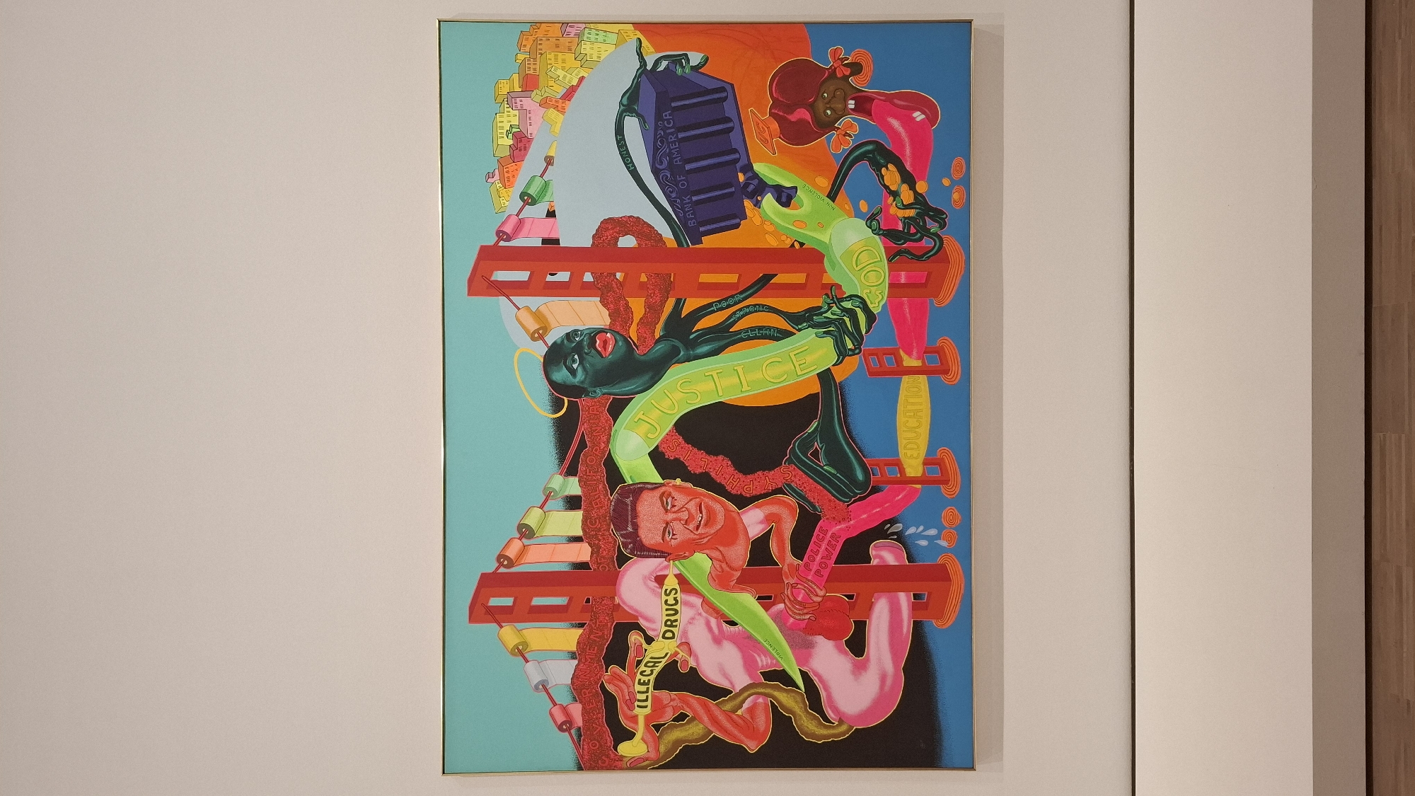 The image shows a framed piece of artwork hanging on a white wall, likely inside an art gallery in Vienna, Austria. The artwork is a vibrant, colorful composition with several surreal and symbolic elements.
A cityscape dominates the top-left corner of the artwork. The central figures include humanoid characters of different colors, intertwined with ladder-like structures. There are serpentine shapes with words written on them, such as "JUSTICE," "EDUCATION," and "ILLEGAL DRUGS." The composition also features symbolic objects, like a Bank of America cash machine.