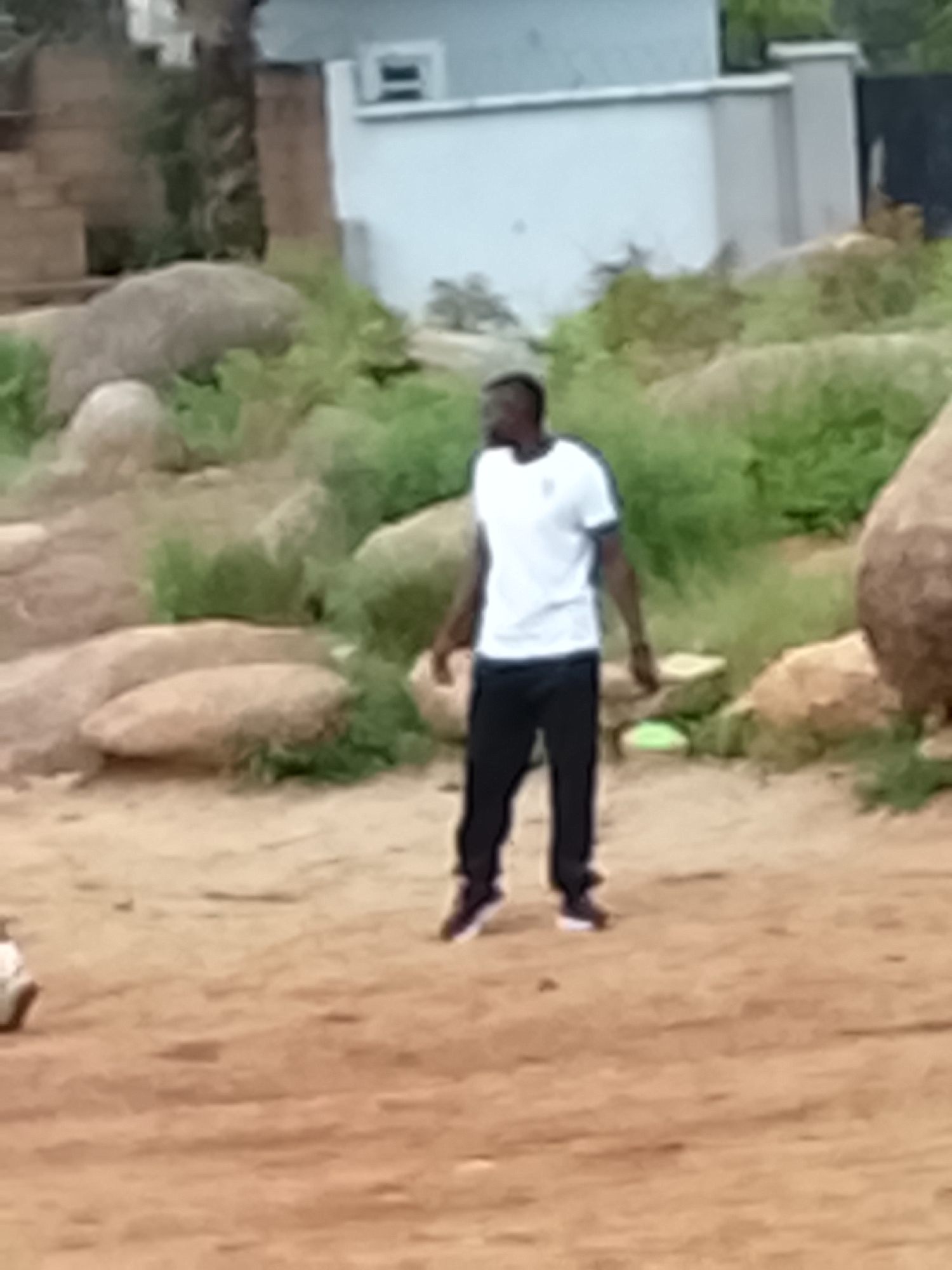 The image shows a man standing in a sandy area with rocks and green vegetation. He is wearing a white t-shirt with dark stripes on the sleeves and black pants. The man is looking to his left. In the background, there is a light-colored building with a fence and some trees. The scene appears to be outdoors in a rural or semi-urban setting. The lighting suggests it is daytime. The specific location in Jos, Nigeria is not identifiable from the image.