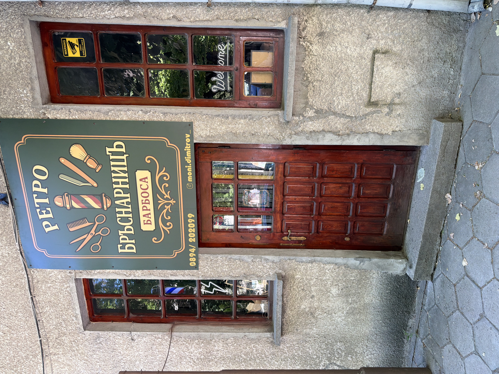 The image shows the entrance to a barbershop named "Retro Barbershop" in Stara Zagora, Bulgaria. The shop's sign, written in Bulgarian, features traditional barber tools. The entrance includes a wooden door flanked by two small, multi-paned windows and a third multi-paned window above the door. One window displays a "Welcome" sign and another displays a "Warning 24hr Surveillance" sign. The building's exterior is a rough, light gray concrete. The entrance has a concrete step leading to a paved walkway with hexagonal concrete blocks.