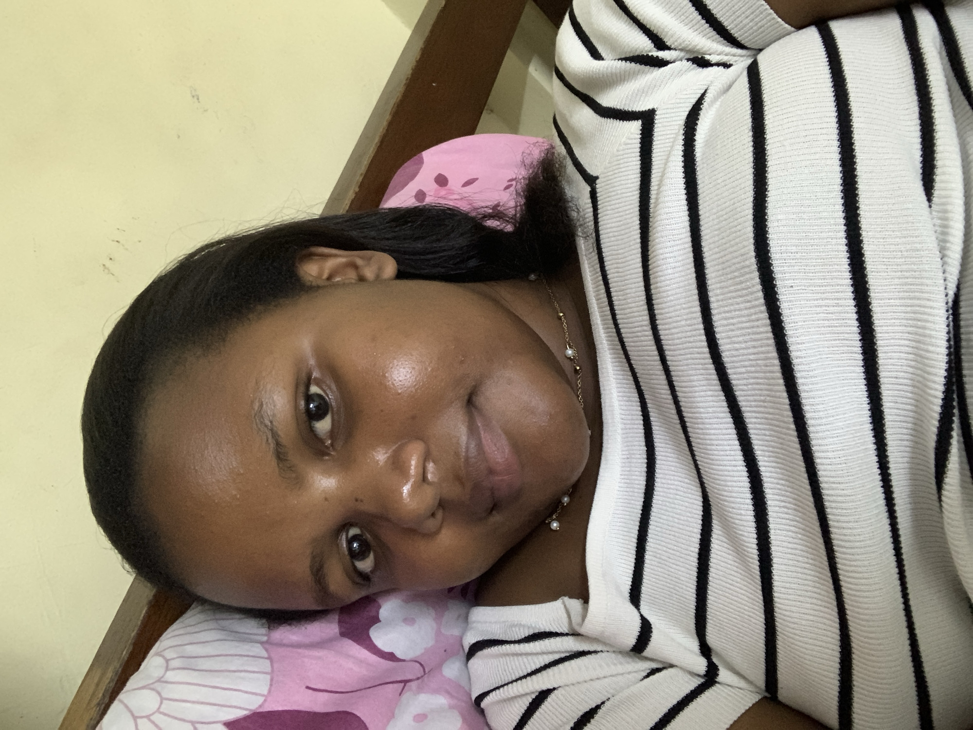 A woman is lying down on a pink floral patterned pillow, wearing a white and black striped shirt. Her head is on the pillow, and she is looking directly at the camera. She has dark hair and brown eyes. A gold necklace is visible around her neck. The background includes a light yellow wall and part of a wooden bed frame. The photograph does not provide enough details to confirm that the location is in the city of Dar es-Salaam, Tanzania. The city and country are not discernible from the visible context.