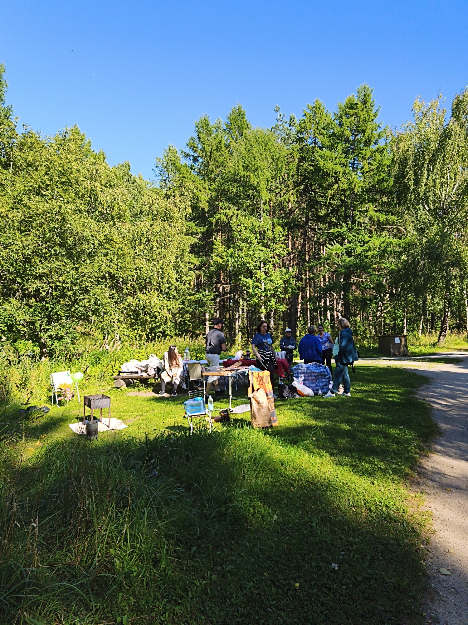 In the city of Maardu, Estonia, a group of people are gathered in a clearing surrounded by a dense forest of deciduous and coniferous trees under a clear blue sky. The scene appears to be a picnic or outdoor gathering. Several folding tables are set up, laden with various items including bags, blankets, and what appears to be food or supplies. People are standing around the tables, some interacting with each other. There are also folding chairs, a barbecue grill, and a couple of portable tables. A dirt path runs along the right side of the clearing, suggesting it might be near a park or recreational area. The ground is covered in green grass and patches of taller weeds. The overall atmosphere suggests a casual, daytime event in a natural setting.