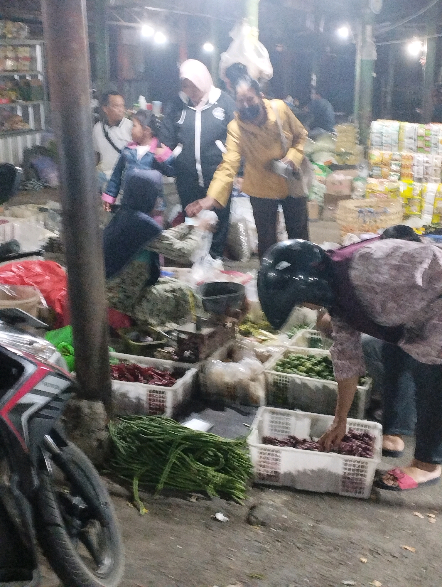 The image depicts a bustling outdoor market scene, likely in Kecamatan Pati, Indonesia, given the context provided. The lighting suggests it is either dusk or an indoor market with artificial lighting. Several individuals are present, engaged in activities related to commerce and shopping.
In the foreground, to the left, a motorcycle is partially visible, indicating a common mode of transport for vendors and shoppers. A person wearing a black helmet and a patterned shirt is bent over a white plastic crate filled with reddish-purple produce, appearing to be selecting items. Beside this crate, another person in blue jeans and pink sandals is also visible.
To the left of the helmeted individual, a large bundle of green string beans lies on the ground. Behind the string beans, a red plastic bag is placed. In the middle ground, various market stalls are set up. One stall displays white crates filled with green vegetables. Another crate contains red chilies. Further back, people in casual attire, including a woman in a light pink headscarf and a black and white jacket, and a person in a yellow shirt, are interacting. The background reveals more market stalls with stacked goods, possibly food items or other merchandise, and the general ambiance of a busy marketplace. The ground surface appears to be dirt or gravel.