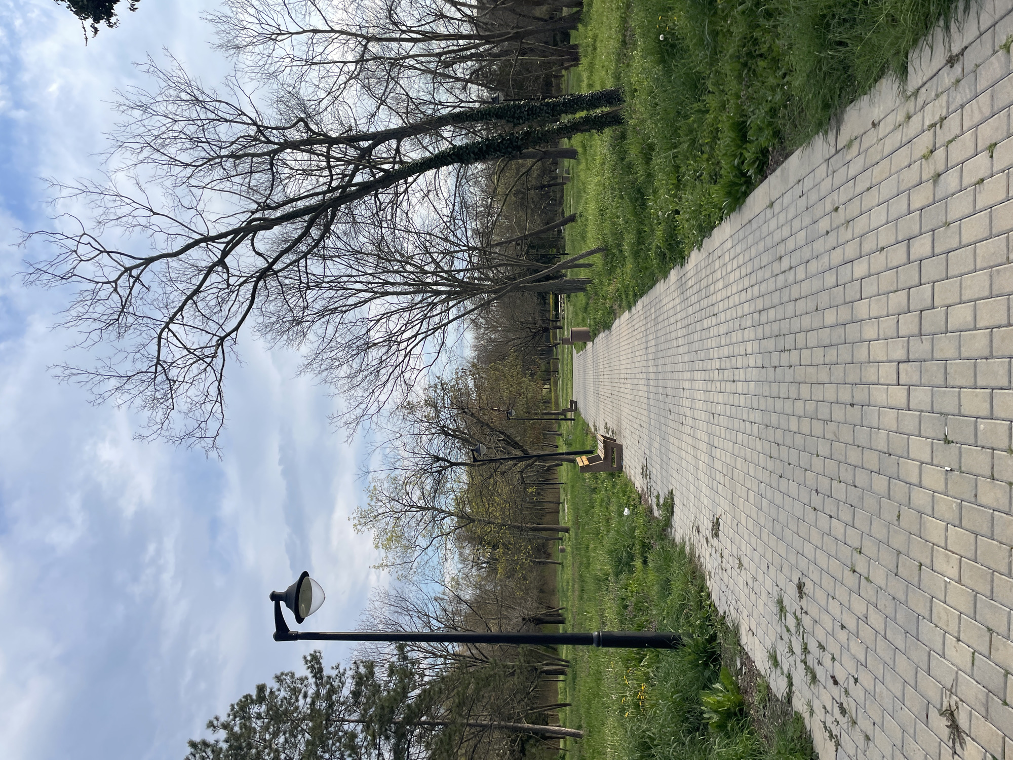 The image depicts a straight, brick-paved walkway in a park setting. The walkway stretches into the distance, bordered on both sides by grassy areas. Trees, some bare and some with early foliage, line the path. A black lamppost stands to the left of the walkway. The sky is visible above the trees, with a mix of blue and clouds. The location is likely a park in Burgas, Bulgaria. There is no people in the scene.