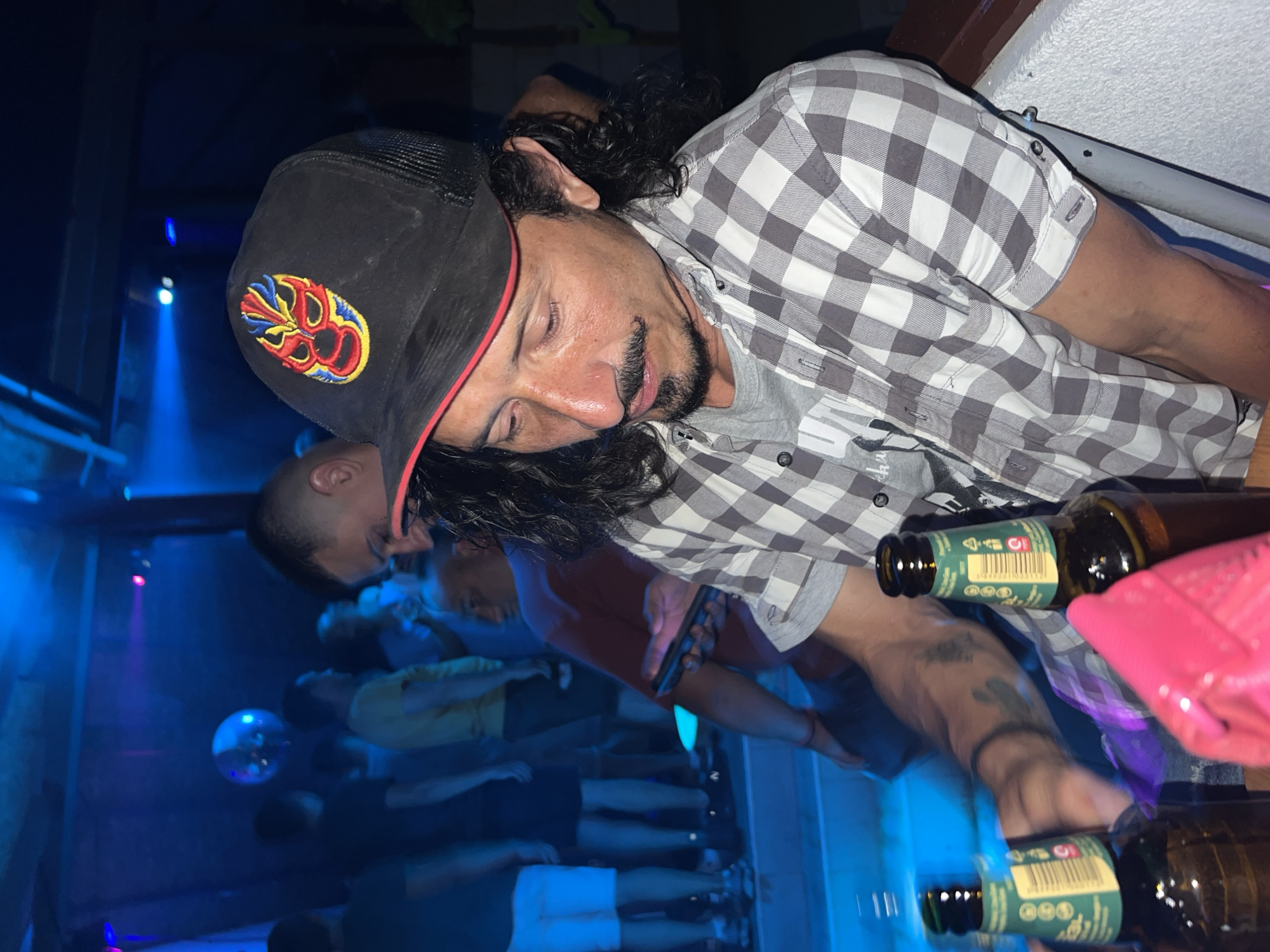 A man wearing a black baseball cap with a colorful luchador mask logo and a grey and white checkered shirt is seated at a table in what appears to be a nightclub or bar. He has dark, curly hair and a mustache and goatee. Two amber-colored beer bottles with green labels are on the table in front of him. A bright pink object, possibly a bag or a piece of clothing, is also on the table. In the background, other people are visible, and the environment is lit by blue and purple lights, with a disco ball in the distance. The scene is likely in Herceg Novi, Montenegro, given the context.
