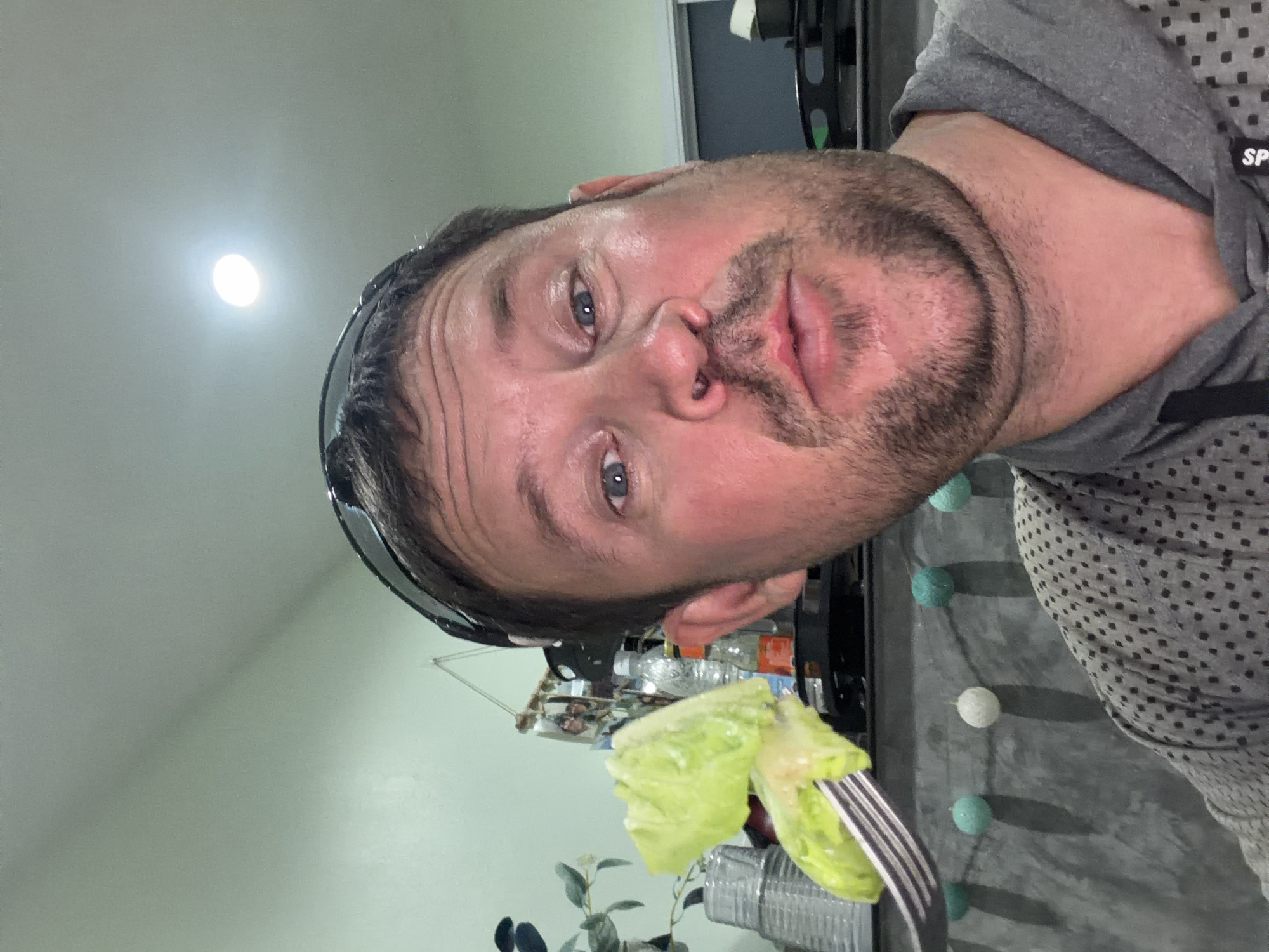 A close-up, selfie-style photograph shows a man in Baan Mae Nam, Thailand, holding a fork with lettuce in front of his face. The man is wearing sunglasses pushed up onto his head and a grey hoodie. He has stubble on his face and his mouth is slightly open. In the background, there is a light green wall, a string of decorative balls, and some bottles on a shelf. The lighting suggests an indoor setting.