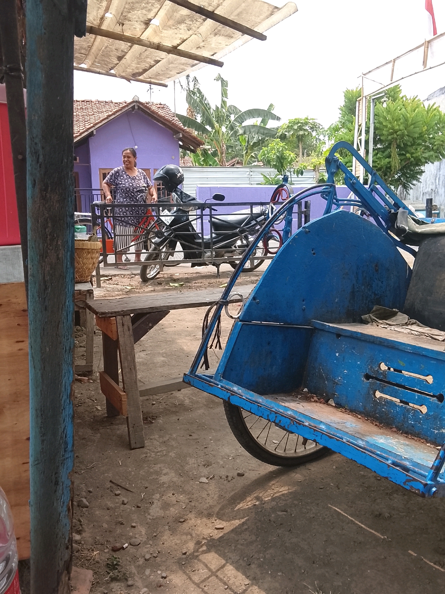 The image depicts an outdoor scene in what appears to be a residential area in Slawi, Indonesia, under a bright, hazy sky. In the foreground, a blue rickshaw, a three-wheeled passenger cart, dominates the right side of the frame. It is weathered and shows signs of age, with peeling blue paint and a worn wooden passenger seat.
Behind the rickshaw, a woman in a patterned blue and white shirt and striped skirt stands near a motorcycle. She appears to be looking towards the left of the frame. The motorcycle has a black helmet resting on its seat. A simple wooden table with visible nails and support beams is positioned between the rickshaw and the woman. A woven basket sits on top of the table.
Further back, a small, light purple house with a tiled roof is visible, surrounded by lush green vegetation, including banana trees. A metal fence runs along the property line. The ground is a mixture of dirt, pebbles, and scattered debris. The overall impression is that of a casual, lived-in environment in a tropical climate.