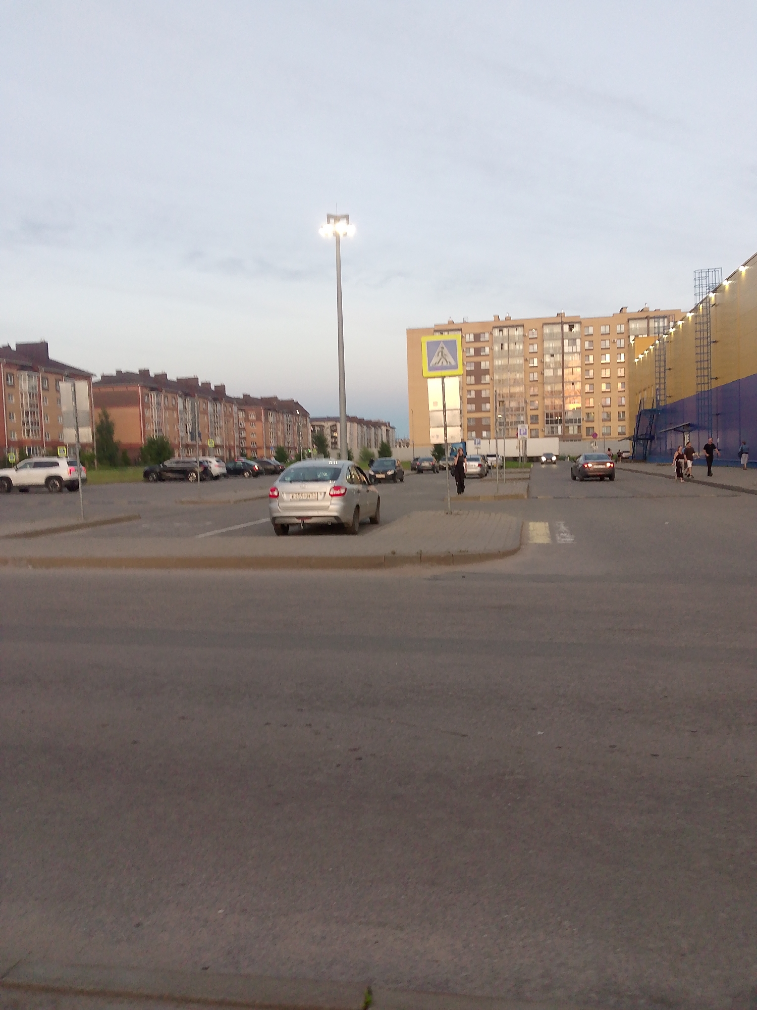 The image depicts a street scene, likely a parking lot, in the city of Shimsk, Russia. In the foreground, a paved road is visible, leading into a parking area. Several cars are parked in the lot, including a silver sedan in the mid-ground. To the left, a white SUV is parked, and further back, a row of apartment buildings lines the street. On the right side of the image, a large, light-colored building, possibly a commercial establishment, is visible. A tall lamppost stands in the middle of the parking lot. The sky is overcast. A few pedestrians can be seen walking in the distance, near the commercial building. The overall scene appears to be a typical urban or suburban environment.