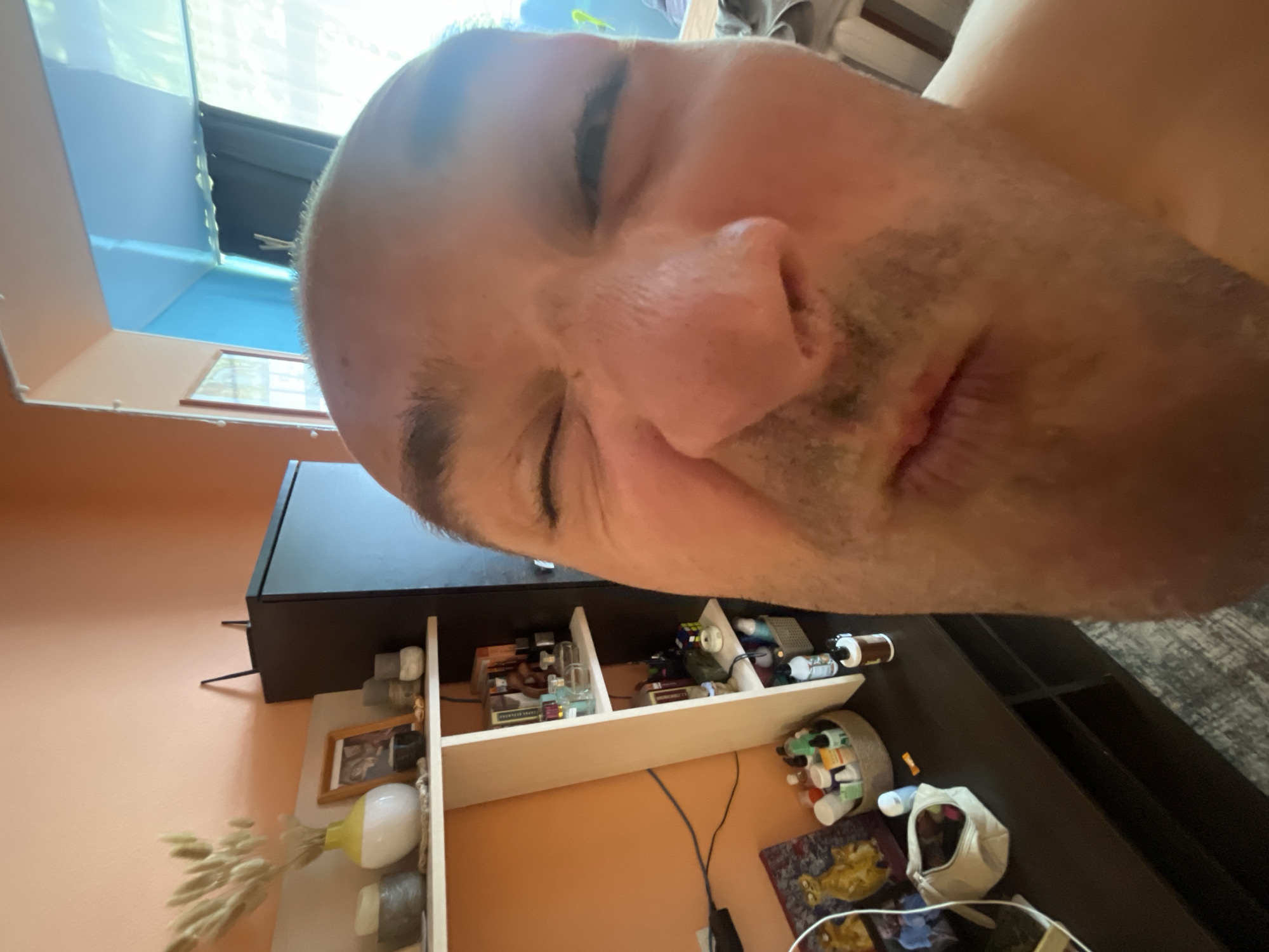 A close-up, low-angle selfie captures a bald man's face, distorted by the wide-angle lens, as he squints with his right eye. The background reveals an interior setting with furniture and personal items. To the left, a peach-colored wall is adorned with shelves holding decorative objects like a yellow and white vase with dried reeds, framed pictures, and small containers. Below the shelves, a dark cabinet displays a basket filled with various toiletries and a white purse. To the right, the scene continues with a blue wall and a window with sheer curtains, suggesting a room in an urban dwelling. The man's expression is playful or awkward, with his mouth slightly ajar.