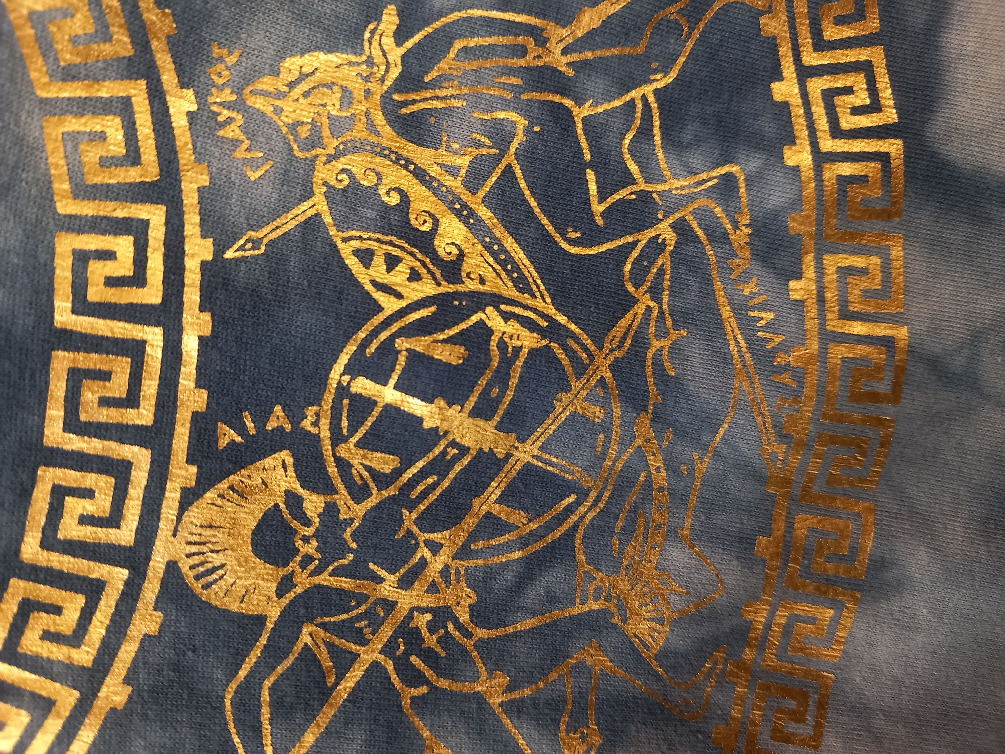 This is a close-up, eye-level, indoor shot of a blue tie-dye fabric with a gold metallic print. The print depicts two figures in combat, rendered in an ancient Greek style. The figures are stylized and appear to be warriors, one with a shield and spear, the other also armed. They are positioned in a dynamic, fighting stance. A decorative Greek key pattern borders the top and bottom of the image. The lighting is even and natural, highlighting the metallic sheen of the gold print. There is no discernible information about the location being in a city.