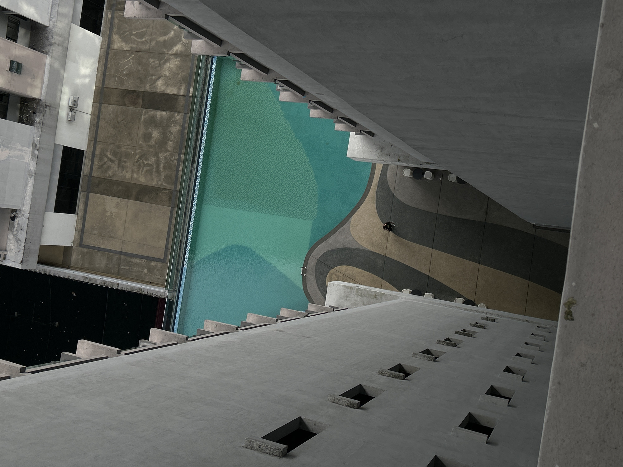 The image presents an angled, high-vantage perspective of a building complex in Kuala Lumpur, Malaysia. The dominant features include stark concrete structures, a turquoise-colored swimming pool with patterned designs, and adjacent buildings.
The right side is dominated by a concrete wall, heavily textured. Below the overhang is a geometric design of rounded stripes. At a distance, near the geometric shapes, is a lone individual.
On the left, the building is multi-storied and of a typical urban design with a black facade. The pool fills a large portion of the central area.
The overall atmosphere is stark and urban.