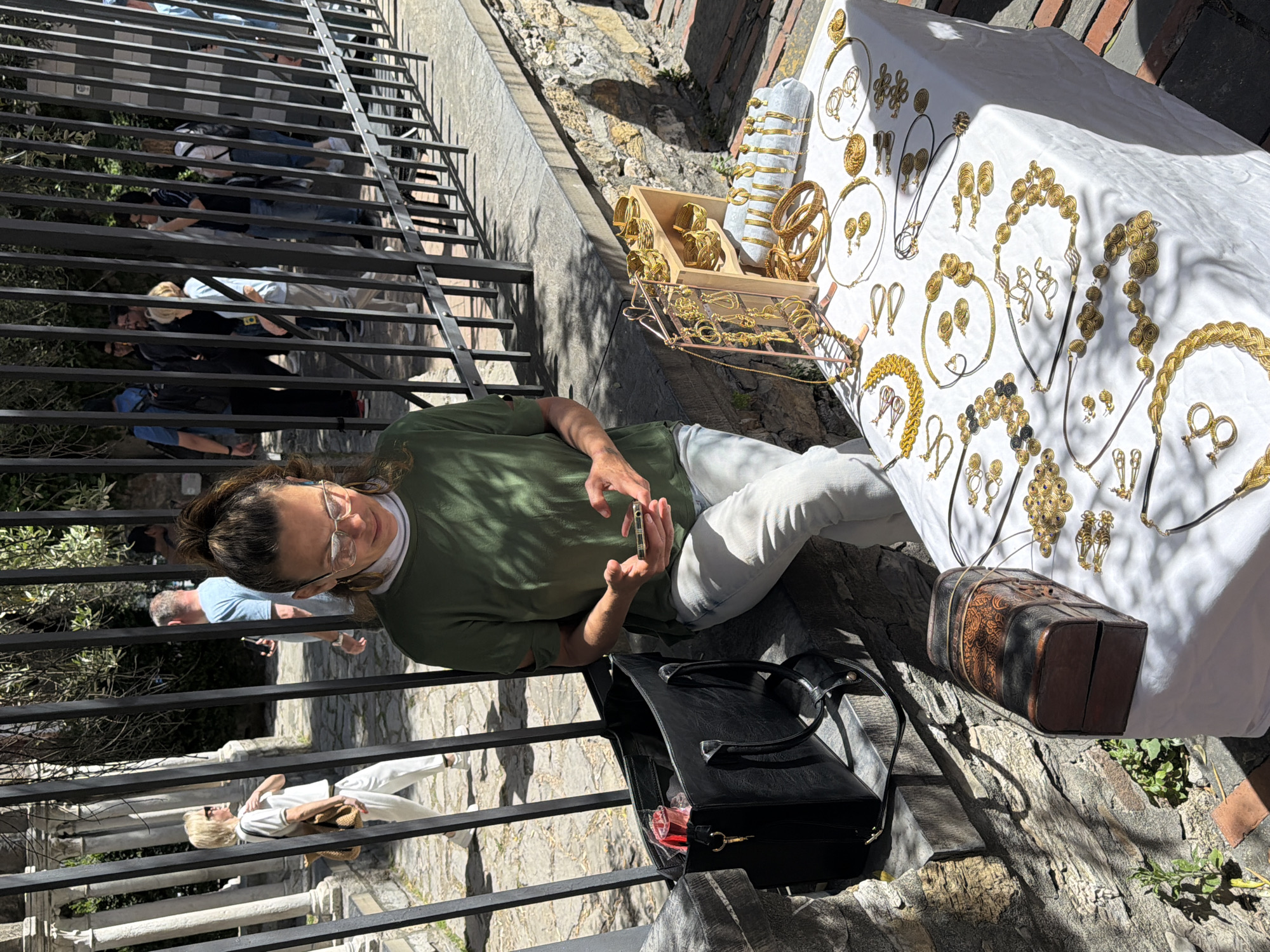 A woman in an olive green shirt and light-colored jeans sits behind a black iron railing on the stone pathway of Salita Santa Maria di Castello, Genoa, Italy. She has brown hair pulled into a ponytail and is wearing glasses. She is holding a cell phone. Next to her is a black leather bag. A white cloth is spread out on a table displaying gold-colored jewelry. A wooden box with more jewelry and a patterned brown suitcase are on the table. Pedestrians are visible behind the railing, walking down the pathway. The environment includes stone structures and stairs.