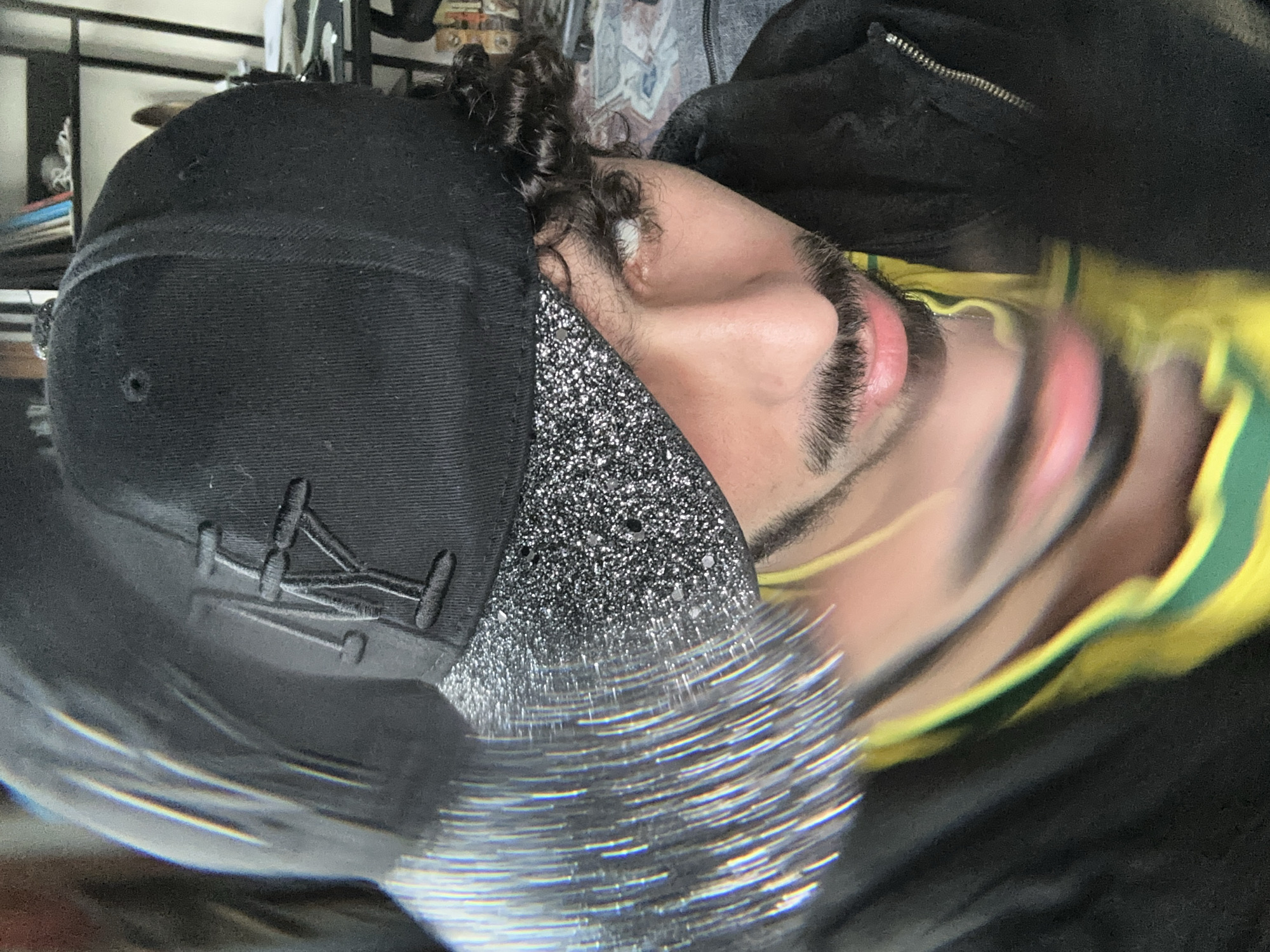 A man is viewed from a close-up, tilted angle, partially obscured by a black baseball cap with an embroidered logo. The cap's brim appears to be covered in a sparkling material, and a blurred, shimmering effect is present near the bottom of the frame. The man's facial features are partially visible; his left eye, eyebrow, eyelashes, part of his nose, and a portion of his mouth are evident. He has dark curly hair peeking out from under the cap. He is wearing a black zippered garment and a garment with yellow and green stripes. The background shows glimpses of shelves and items suggesting an interior setting. The location, Palaiseau, France, is not evident in the image.
