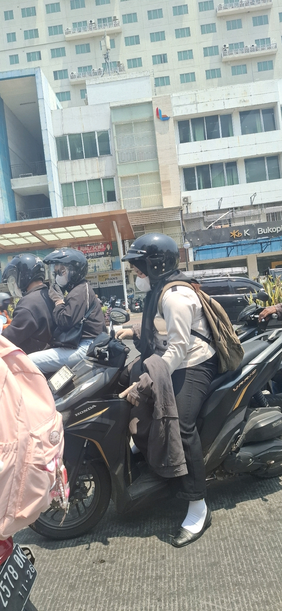 A group of people are on motorcycles in an urban environment, likely in Surakarta, Indonesia. In the foreground, a person wearing a black helmet, face mask, white long-sleeved shirt, and black pants is seated on a dark-colored motorcycle. They have a beige backpack slung over their left shoulder and are holding a dark jacket. To the left of this person, another individual with a black helmet and face mask is visible, also on a motorcycle. Further to the left, a person wearing a light pink backpack and partially visible clothing is positioned. The background shows multi-story buildings, with the closest one featuring large windows and a signage with red and blue abstract logo and "Bukopin" text, suggesting a commercial or financial institution. The motorcycles appear to be parked or moving slowly on a paved road with visible markings. The lighting suggests it is daytime with bright sunlight.