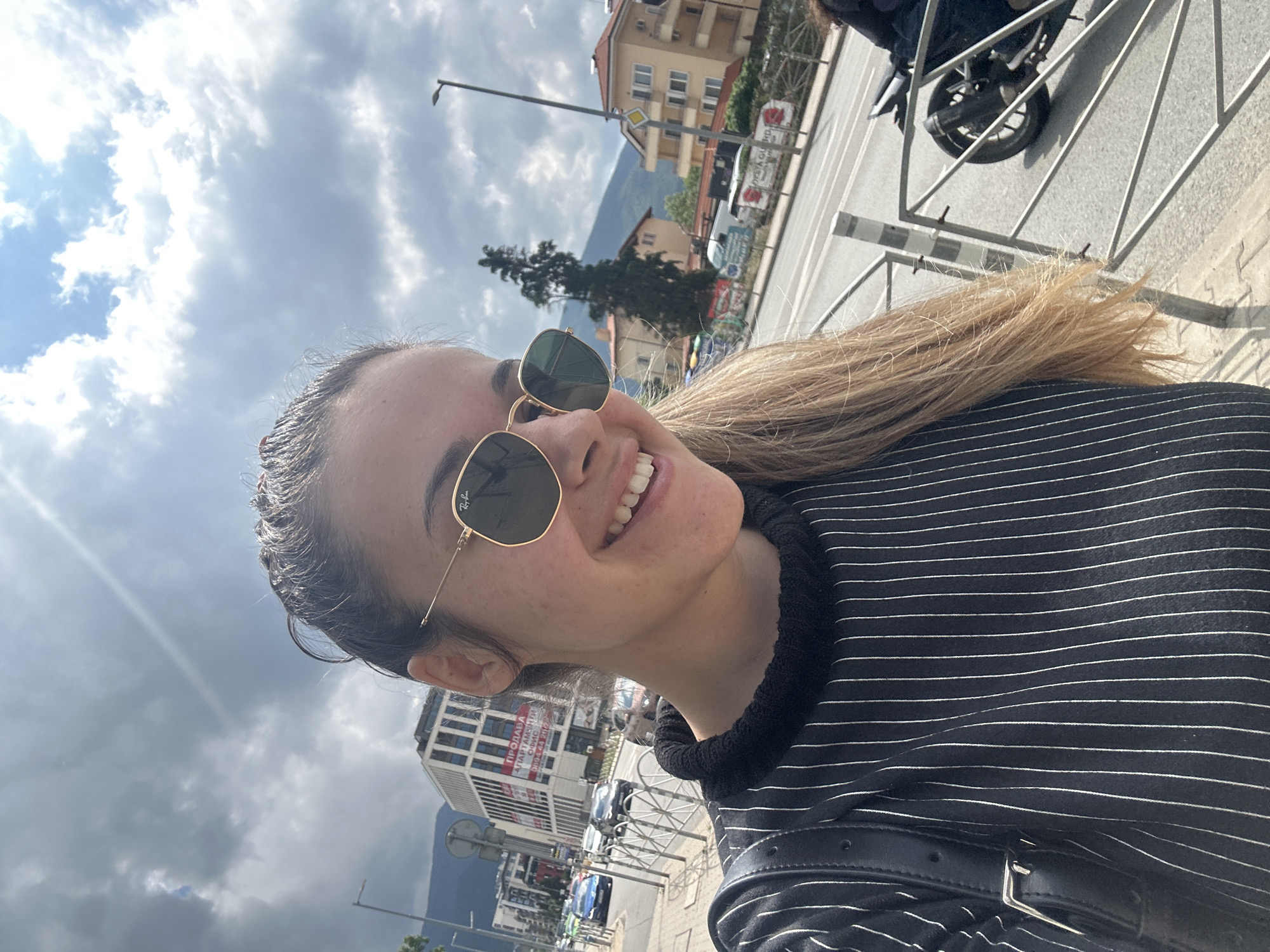 This dynamic, obliquely angled image captures a smiling young woman against an urban backdrop in Sofia, Bulgaria. The subject, positioned in the foreground, is a woman with light brown hair pulled back, wearing gold-framed Ray-Ban sunglasses. She is dressed in a black top with thin white horizontal stripes and a black shoulder bag with a silver buckle is visible. Her expression is one of broad happiness, indicated by her wide smile showing white teeth.
The setting is a city street, with various multi-story buildings, including both older residential structures with balconies and a more modern white building. In the distance, mountains are discernible, characteristic of Sofia's geography near Vitosha. The street features a light pole, a metal fence, and parked cars, with a motorcycle partially visible near the fence. Trees are scattered throughout the scene.
The photograph appears to be a casual selfie or close-up portrait taken during the day. The bright sunlight, casting some glare and visible shadows, suggests it is a clear day, though there are scattered white and grey clouds in the blue sky. The overall mood conveyed is cheerful and relaxed.
Notable text details include "Ray-Ban" visible on the left lens of the sunglasses. On a prominent white building in the mid-ground, Cyrillic text reads "ПРОДАВА" (PRODAVA - FOR SALE) and "АПАРТАМЕНТИ" (APARTAMENTI - APARTMENTS), confirming the Bulgarian context of the location.