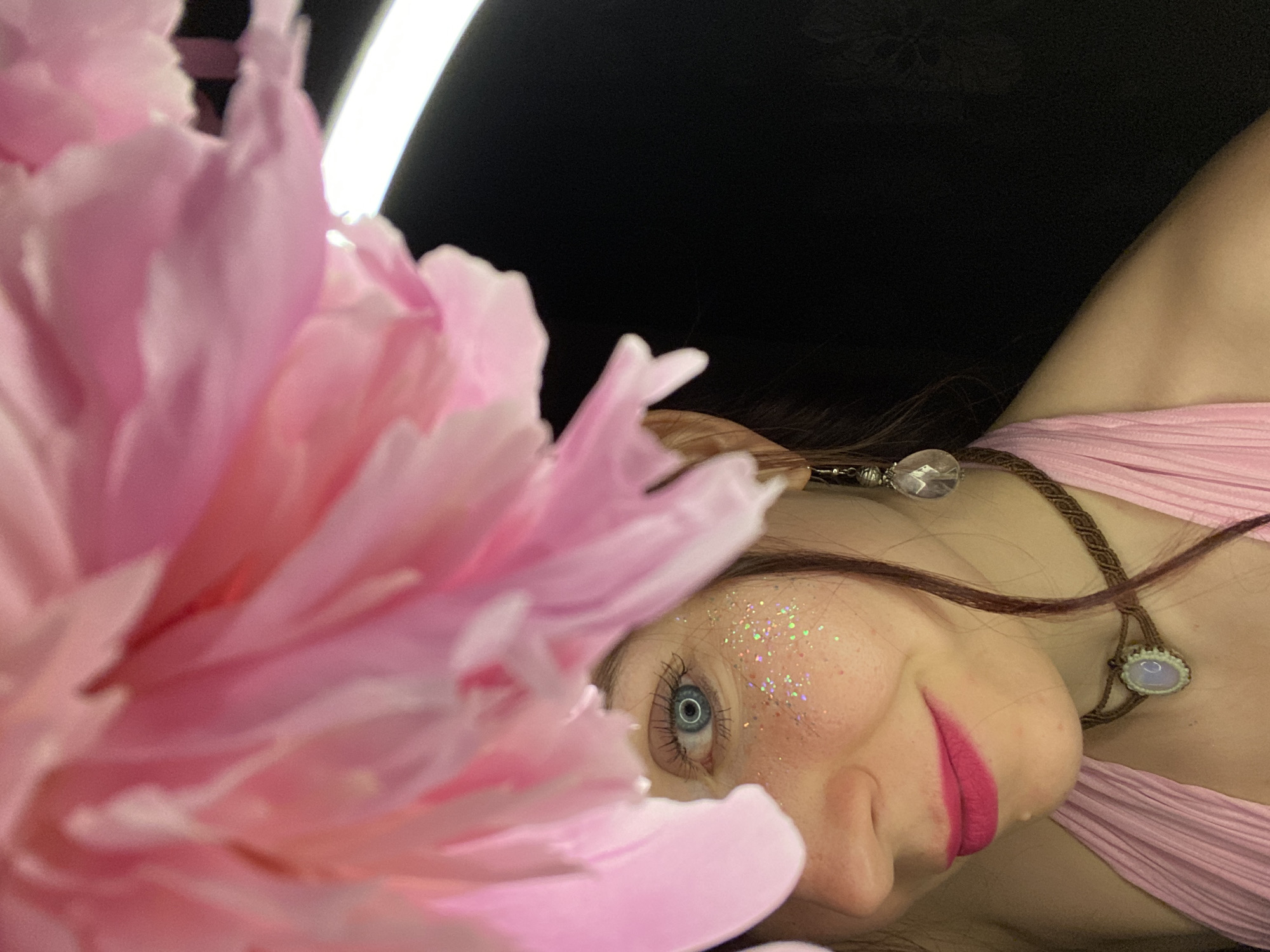 A close-up, artistic portrait captures a person, likely female, lying down with their head tilted, partially obscured by a large, soft pink flower, possibly a peony, in the foreground. The person has a striking blue eye, long eyelashes, and wears vibrant pink lipstick. Glitter is artfully applied to their cheek and temple area, creating a whimsical or fairy-like aesthetic. Long, dark brown hair frames their face. They are adorned with a braided brown necklace featuring two distinct pendants: one light-colored, possibly iridescent, and another clear, faceted one near the ear. A pink pleated garment is visible on their shoulder and chest. The background is predominantly dark, with a bright white light source visible in the upper left portion of the image. The overall setting appears to be an indoor or studio environment, using controlled lighting to achieve a dramatic and ethereal mood. The person maintains a subtle, gentle expression. No visible text is present in the image. The location context for this media file is Chaikovsky, Russia.