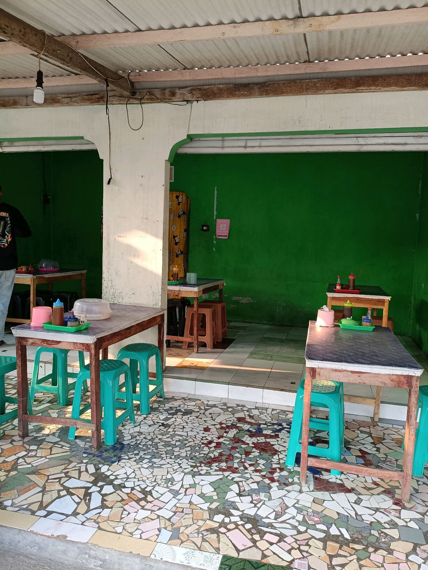 The image shows an outdoor seating area of a restaurant in Maguwoharjo, Indonesia. The area is covered by a corrugated metal roof supported by wooden beams. The walls are painted bright green, and the floor is tiled with a mosaic of broken ceramic pieces.
There are several tables and chairs set up for dining. In the foreground, a rectangular table with a dark, possibly laminate, top is surrounded by teal plastic stools. On the table, a green tray holds a white plastic container with a lid, a bottle of brown sauce, and a small pink container. Another similar table with teal stools is visible further back.
In the middle ground, a person wearing a black t-shirt with a graphic design and blue jeans stands with their back to the camera, facing a table set up against the green wall. This table also has a dark top and is equipped with a pink container and a bottle of sauce. Further to the right, another table with a wooden frame and dark top is set with a red tray containing two bottles, one red and one brown. This table is also surrounded by brown plastic stools.
The environment appears to be a simple, open-air eatery. The lighting suggests it is daytime, with sunlight casting shadows on the floor and walls. The overall scene depicts a casual dining setting, likely a local restaurant.