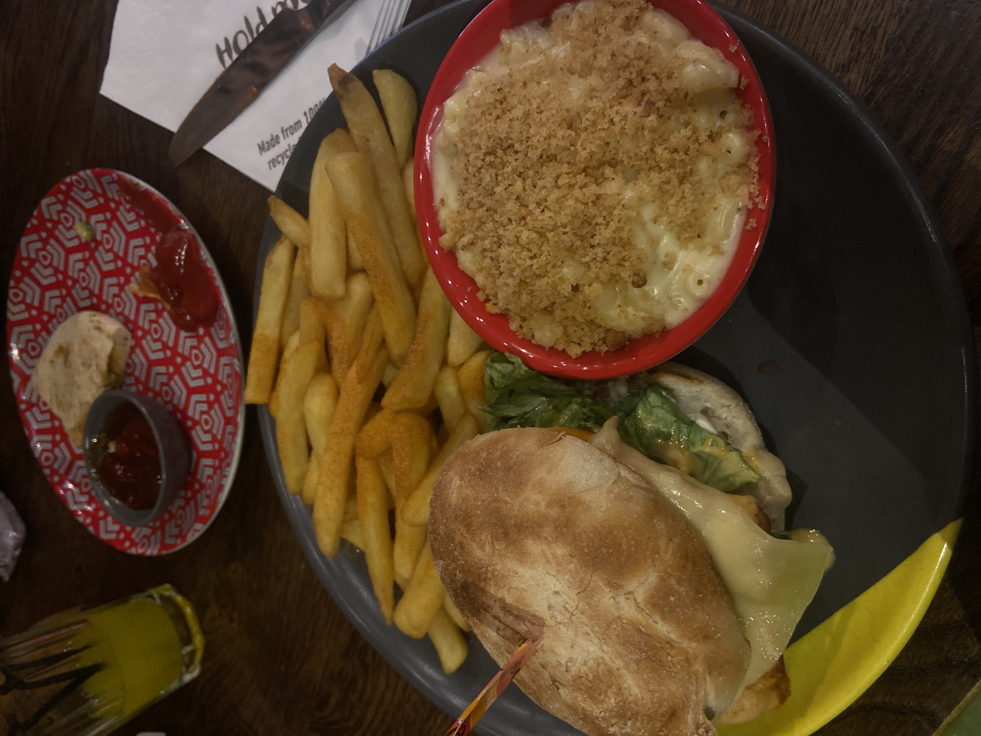 The image shows a meal on a dark wooden table. A dark grey plate contains a burger, fries, and a red bowl filled with macaroni and cheese topped with breadcrumbs. A small, patterned red plate holds a small dish of red sauce and some bread. A glass containing a yellow liquid is also visible. A napkin and cutlery are situated near the plate. It is impossible to tell if it is in London.