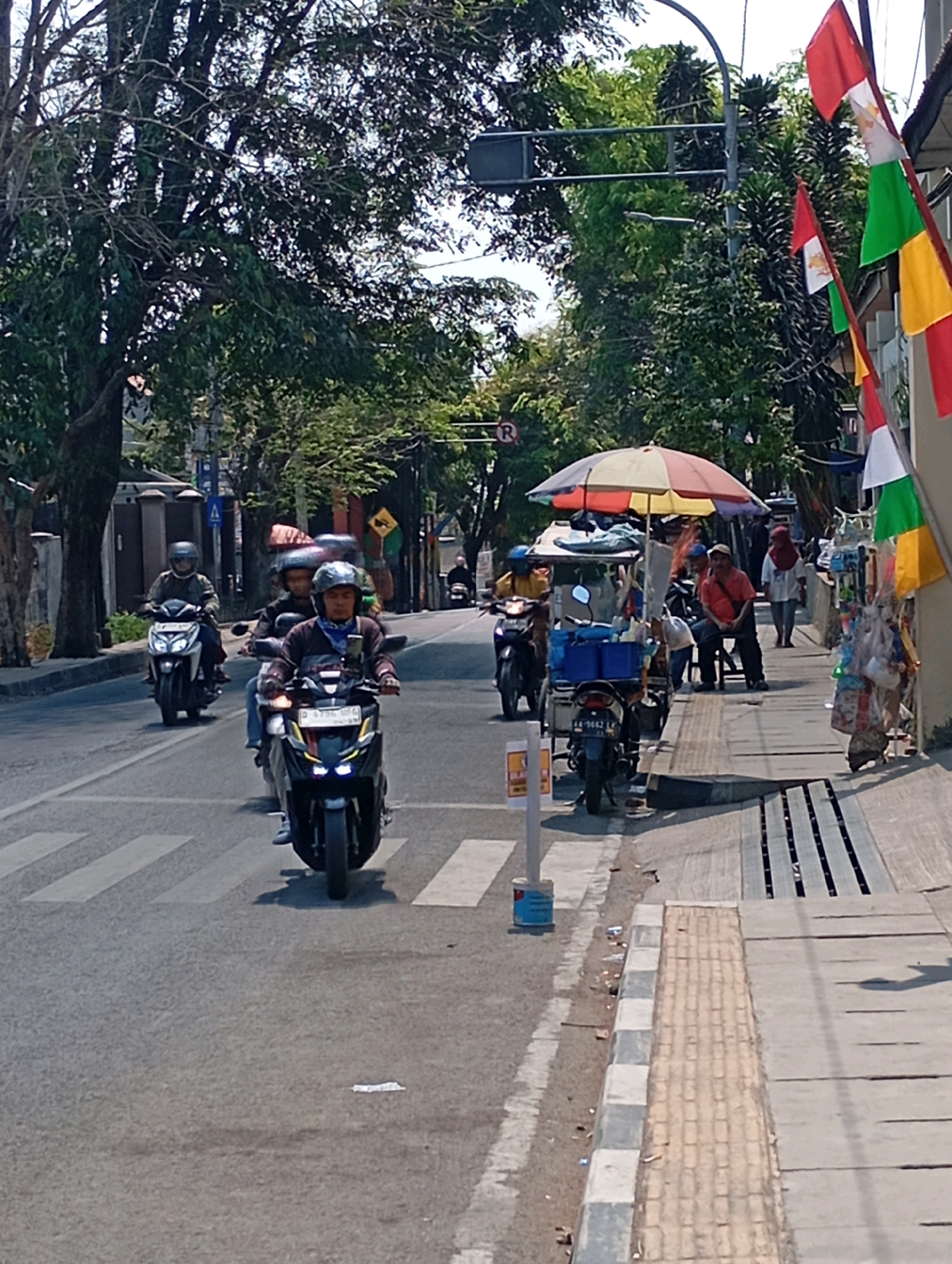 The image depicts a street scene in Cimahi, Indonesia, with several motorcycles on the road and pedestrians on the sidewalk. A street vendor with a colorful umbrella is visible on the sidewalk, alongside a cart. Flags, possibly national or celebratory, are hanging from a building on the right side of the image. Trees line the street, providing shade. Traffic signs, including a circular sign with a red border and an "R" inside, are present. The road appears to be asphalt, with a pedestrian crossing marked by white stripes. The overall atmosphere suggests a typical urban environment with active street life.