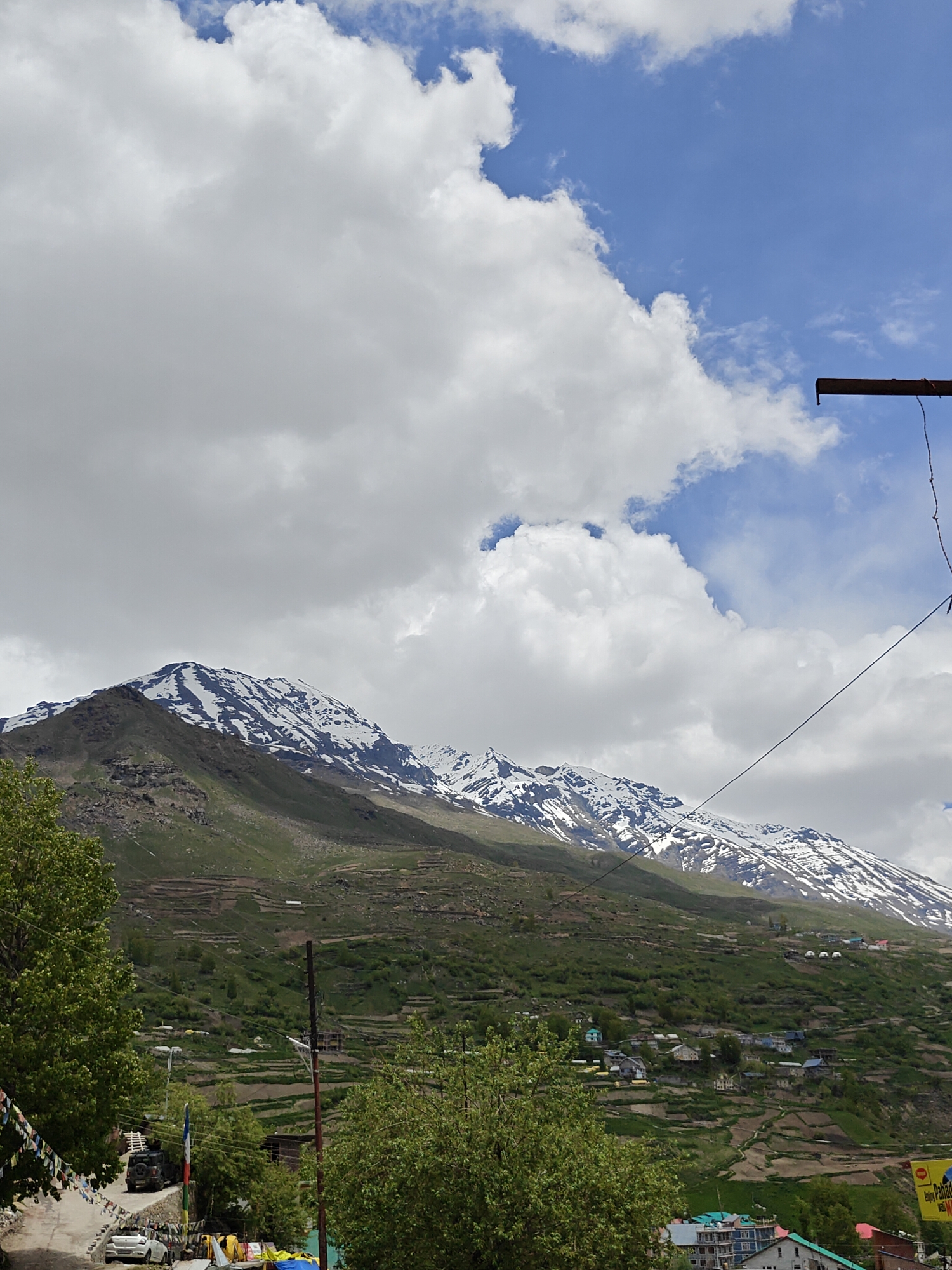 The image captures a mountainous landscape in Yamling, India, under a partly cloudy sky. The foreground features a street scene with parked cars and sparse vegetation. Multi-colored prayer flags are visible along the road. In the midground, a large mountain covered with green vegetation and patches of snow dominates the view. The terraced slopes of the mountain accommodate scattered houses and buildings. In the background, the sky is a mix of blue and white clouds. A utility pole and wires are present in the upper right corner of the frame, intersecting the sky and adding a human element to the natural landscape.
