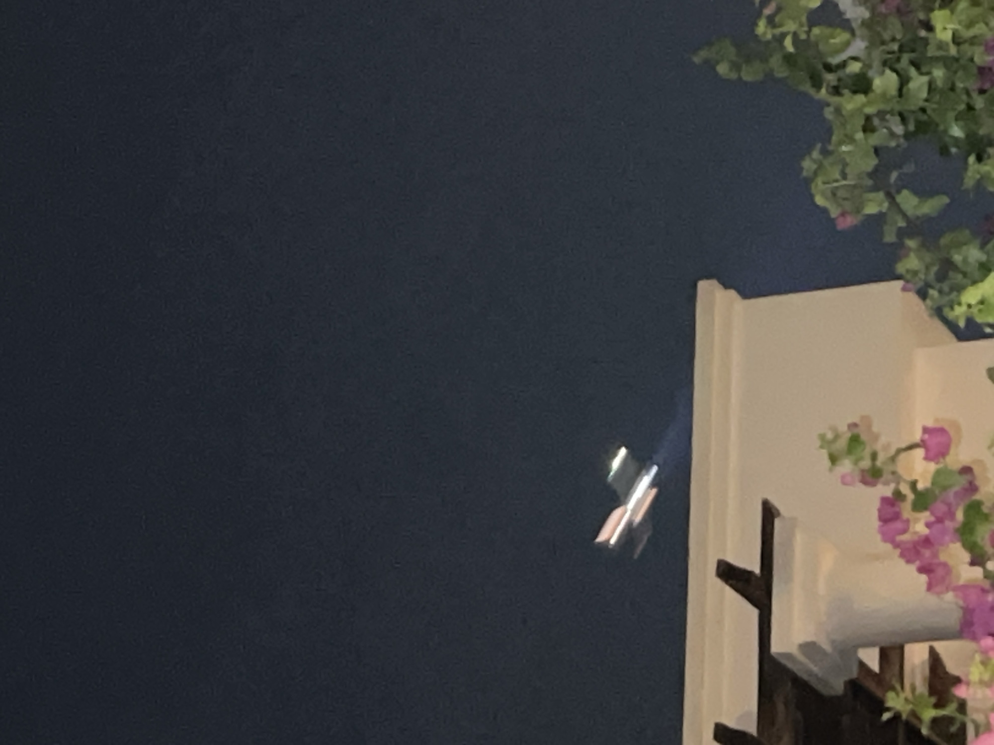 The image, taken at night in Sharm El Sheikh, Egypt, shows a blurry airplane in flight against a dark blue sky. The airplane appears to be illuminated, with streaks of light suggesting its movement. Below the sky, the top of a white building with decorative wooden beams is visible, and a cluster of pink and purple flowers, likely bougainvillea, is in the foreground.