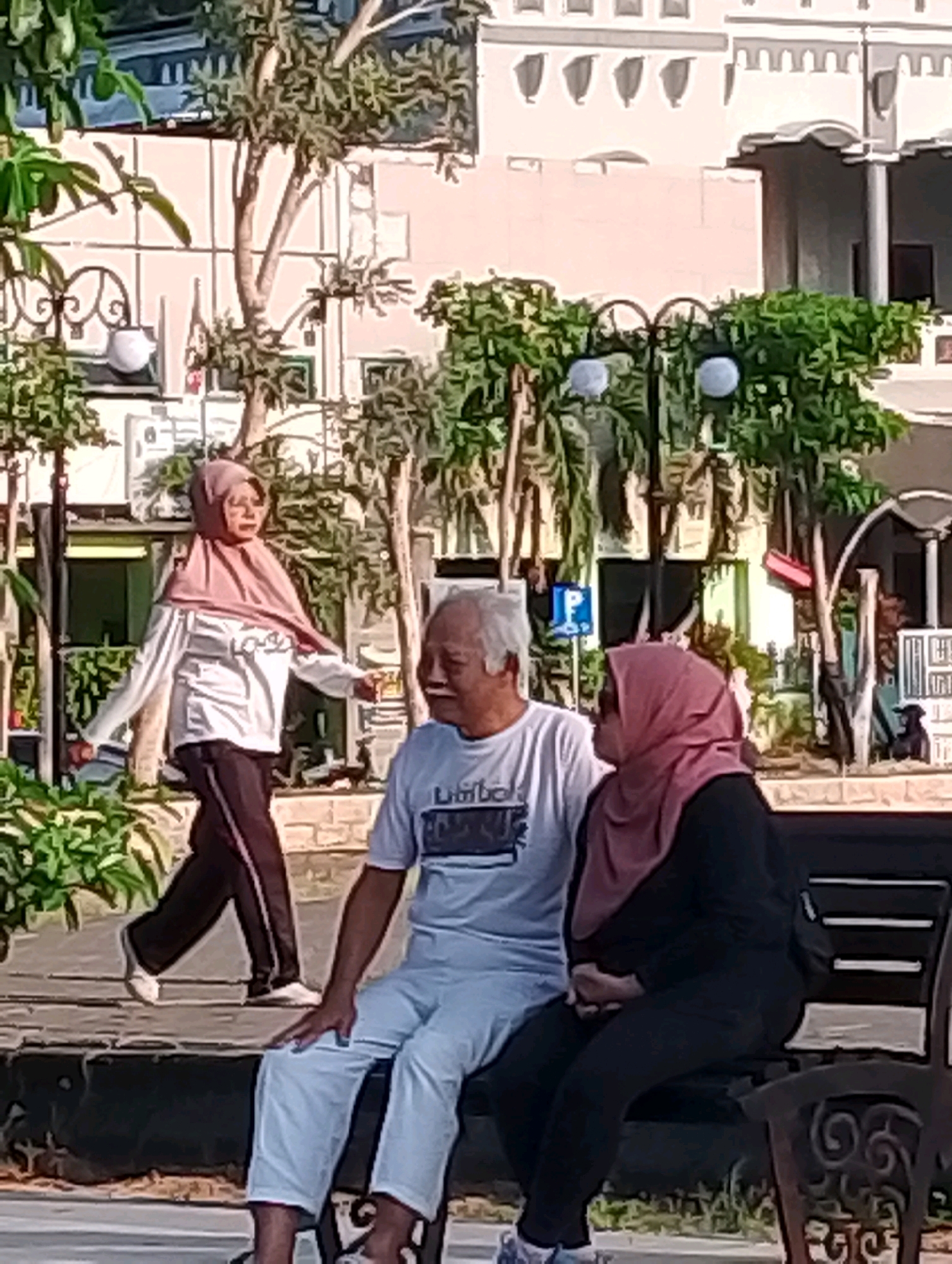 The image depicts an outdoor scene in Probolinggo, Indonesia, likely a public park or promenade, with a building facade and trees in the background. In the foreground, a man and a woman, both wearing hijabs and modest athletic wear, are seated on a dark-colored bench. The woman is on the right, dressed in black, with her hands clasped in her lap. The man, to her left, wears a white t-shirt and light blue pants. His left hand rests on his knee, and he is looking towards the woman, appearing to be engaged in conversation.
To the left of the seated couple, a woman in a light pink hijab, white long-sleeved shirt, and dark pants is walking away from the camera. She is captured mid-stride, with her left arm bent and her right arm extended slightly. Green foliage is visible in the lower left corner of the frame, and a lamppost with decorative elements is positioned between the walking woman and the seated couple. A blue parking sign is also visible in the mid-ground behind the seated man. The overall atmosphere suggests a casual, daytime outing in a city environment.