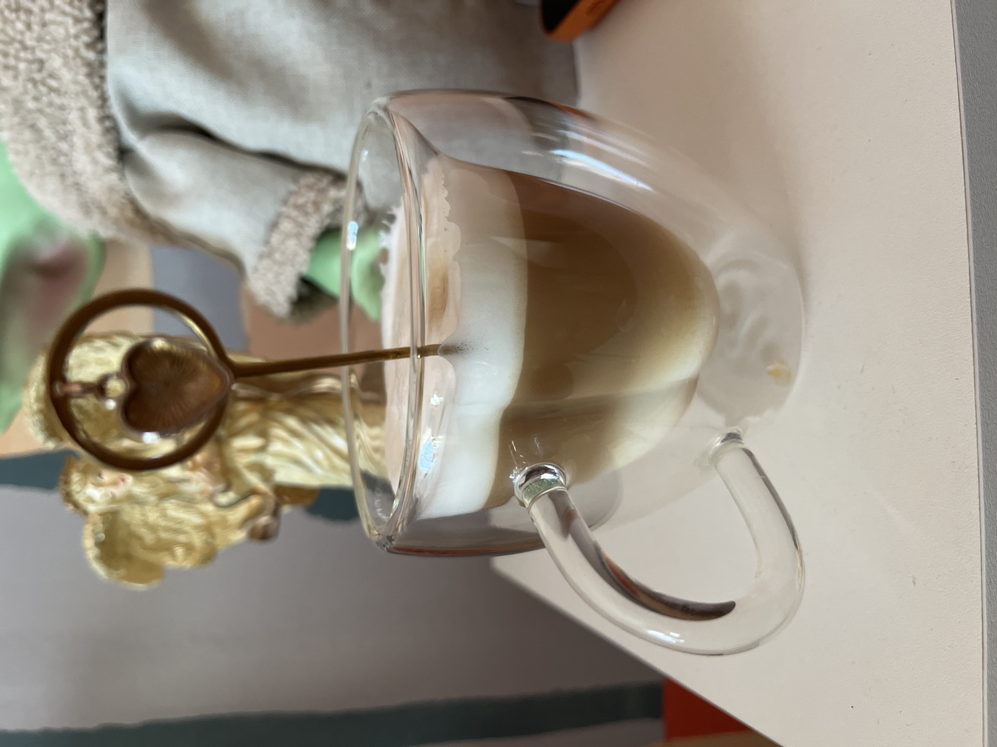 A tilted view shows a clear, double-walled glass mug filled with a beverage, possibly a latte, featuring a foamy top and a visible coffee layer. A gold-colored spoon with a heart-shaped ornament at the top protrudes from the foam. The mug rests on a light-colored surface. Part of a furry, light-colored object and a golden-colored object are visible behind the mug. The location is unknown, and there is no indication it is in Chaikovsky, Russia.