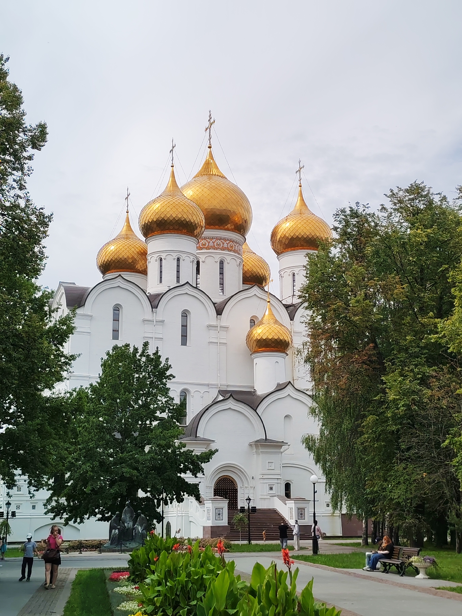 The image depicts a bright white cathedral with multiple golden domes, set against a cloudy sky. The cathedral features traditional Russian Orthodox architecture, with arched windows and decorative friezes. The foreground is a well-maintained park with lush green trees and flowerbeds. Several people are visible: a woman in a pink top and dark skirt walks on a paved path, a young boy in a blue shirt and dark pants walks beside her, and another woman sits on a park bench to the right, looking down at her phone. In the distance, near the cathedral entrance, a few individuals are standing or walking up stairs. The overall scene suggests a peaceful, public space with both religious and recreational elements. The location is identified as the Assumption Cathedral in the city of Rusanovo, Russia.