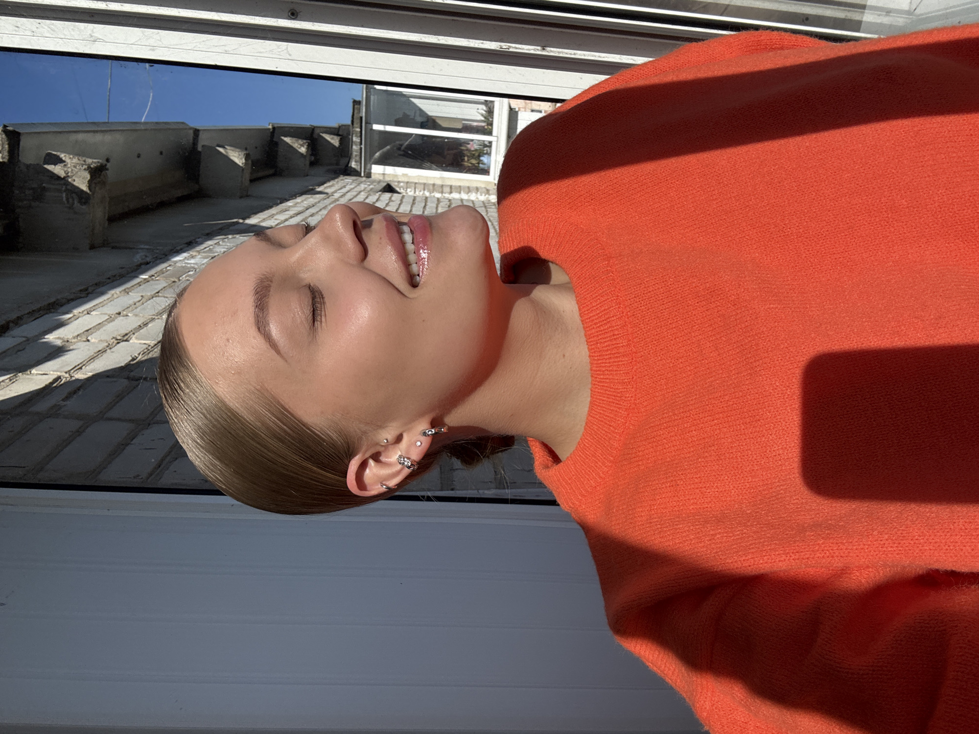 A woman with blonde hair pulled back is smiling upward toward the sun and a light blue sky. She has multiple earrings in her left ear. She is wearing an orange sweater. The woman is lying down or leaning back near a structure with white siding or a window. Beyond the siding is a low building with concrete supports. Based on the presence of Cyrillic signage and architectural styles typical of Russia, the location may be Chaikovsky, Russia, though this cannot be confirmed conclusively from the image alone.