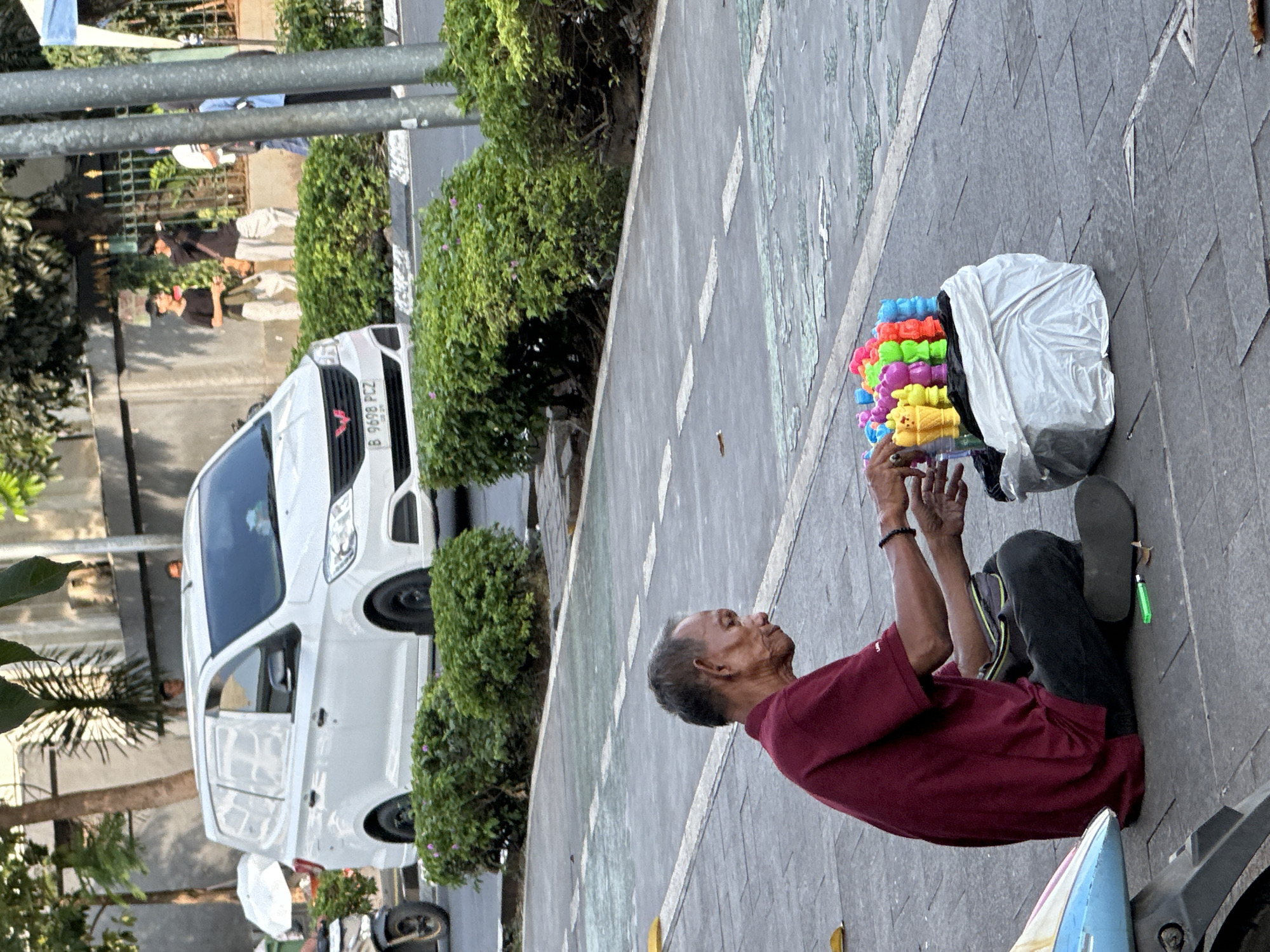An elderly Asian man is kneeling on a paved sidewalk in West Jakarta, Indonesia. He is wearing a maroon shirt and dark pants, and has one sandal on. A white plastic bag containing colorful plastic toys sits to his left. He is looking up and holding his hands up in front of the bag. Behind him, a white van is parked on the street, next to a row of green bushes. Pedestrians can be seen in the background. The license plate on the van reads "B 9698 PCZ".