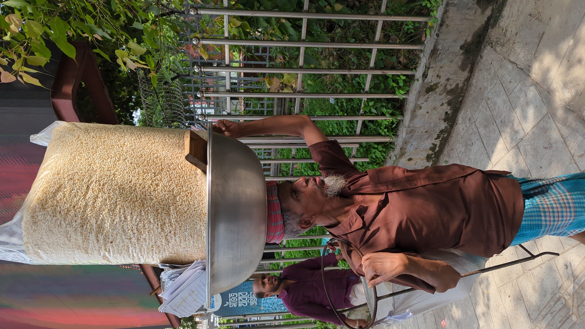 An elderly man with a gray beard wearing a brown shirt, a pink and red head wrap, and a blue and white checkered lungi is balancing a large metal bowl on his head. The bowl contains a large bag of puffed rice, which sits atop a wooden block. The man is holding onto the bowl with his right hand for balance. In the background, there is another man with a beard wearing a purple shirt. The scene is set on a sidewalk in Dhaka, Bangladesh, near a metal fence and some greenery. A colorful advertisement or sign is visible in the lower left corner.