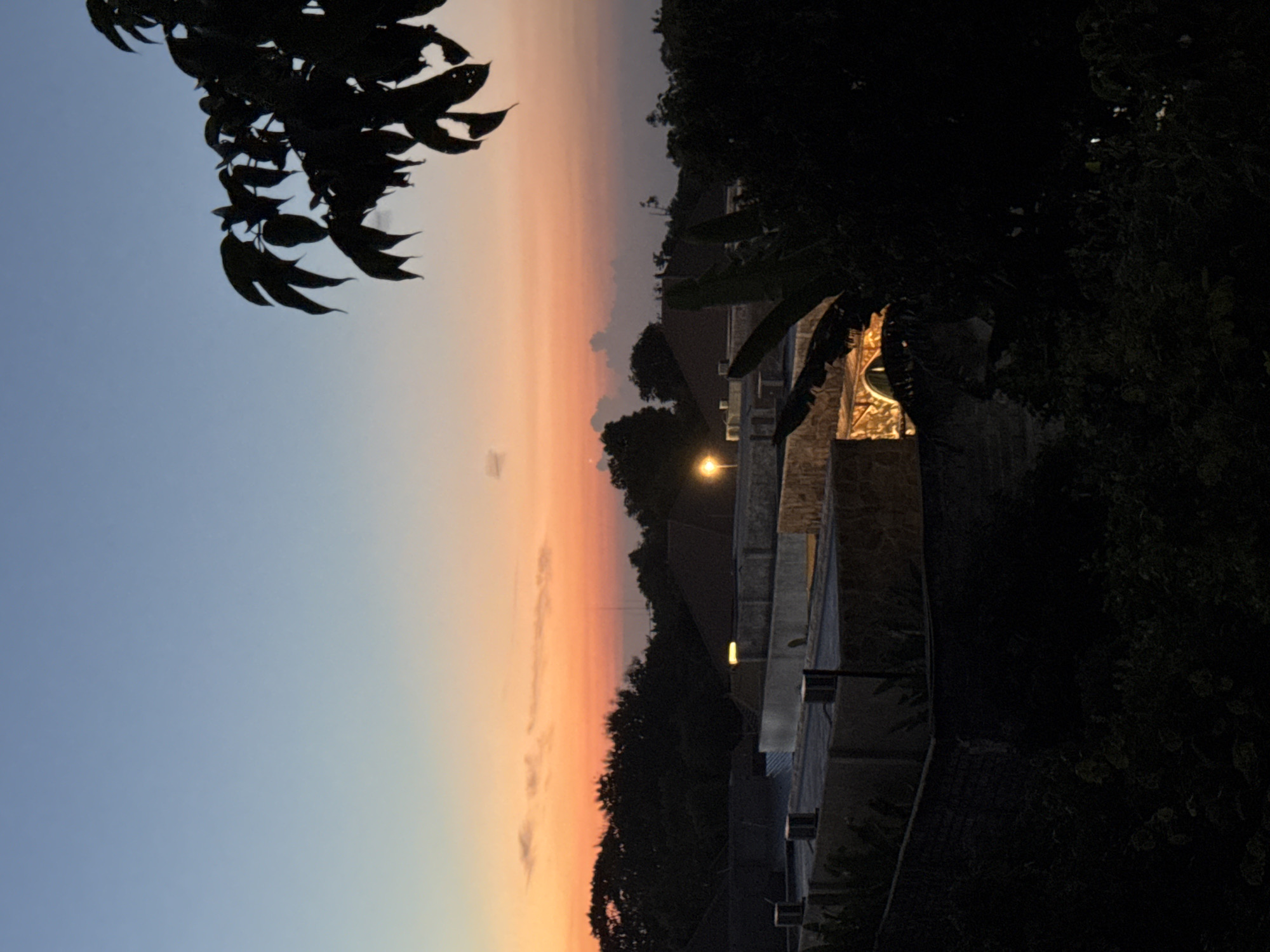 This media file captures a tranquil twilight scene in Kuta Selatan, Indonesia, depicting the transition from day to night. The sky, occupying the upper and central parts of the frame, displays a soft gradient of colors. It transitions from a clear, light blue-grey in the upper left to warmer hues of orange, pink, and yellow towards the right and lower horizon, characteristic of a sunset or dusk. Subtle, wispy clouds are visible within the warmer sections of the sky.
In the foreground, dark, silhouetted tree branches with prominent leaves extend from the upper left corner, partially framing the expansive sky. A dense line of dark trees and foliage forms a natural, undulating horizon line across the lower half of the image, separating the sky from the built environment.
Below the treeline, elements of human construction emerge from the deepening shadows. Structures, possibly parts of buildings or walls, are visible, mostly rendered in silhouette. Artificial light sources punctuate the scene; a bright, yellow-glowing street lamp is prominent towards the middle-right. Further to the right, a section of a building appears illuminated with warm yellow light emanating from windows or interior spaces, creating a welcoming glow. Additional smaller, square-shaped lights are affixed to a wall or railing in the lower right, also emitting a soft, warm light. There is no discernible human activity or visible text in the image. The overall atmosphere is peaceful and serene.