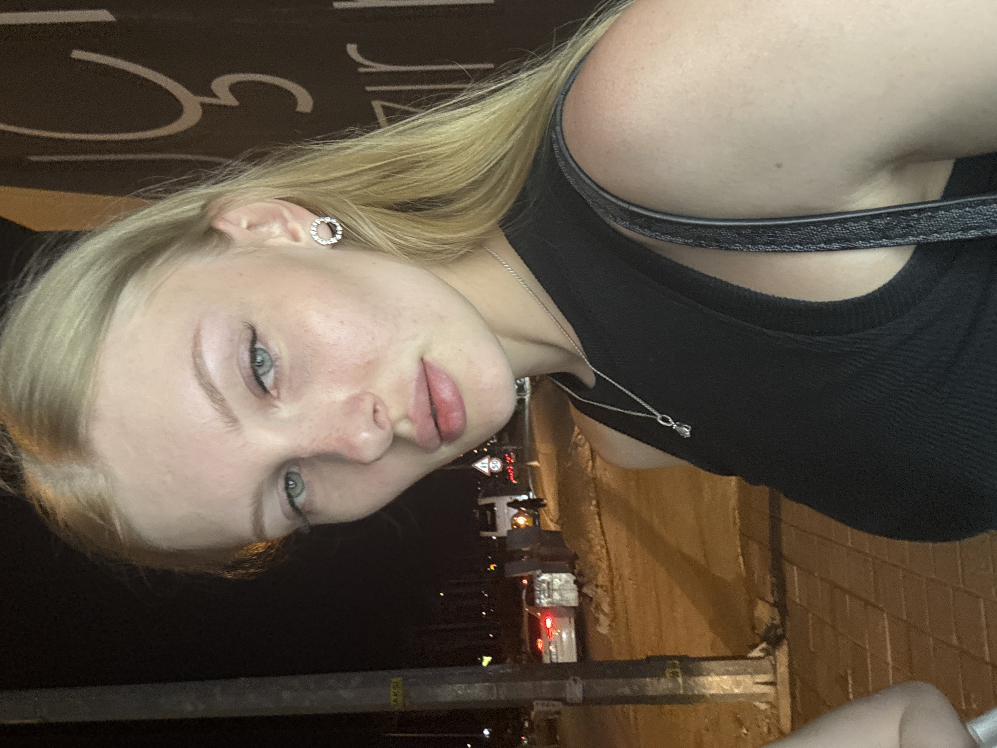A full-frame, close-up, slightly high-angle, night-time, outdoor, selfie of a young Caucasian woman with blonde hair and blue eyes. She is wearing a black tank top, a silver necklace with a pendant, and small hoop earrings with clear stones. Her hair is long and falls over her right shoulder. Her skin is fair with a few freckles. The background includes a dimly lit street scene with a sidewalk, a traffic pole with signage, a car with its taillights on, and blurred figures and vehicles in the distance. There is signage on a dark wall to the right of the woman that appears to be in Turkish. The woman is looking towards the camera with her head tilted slightly. The scene is likely in Kepez, Turkey, based on the Turkish signage.