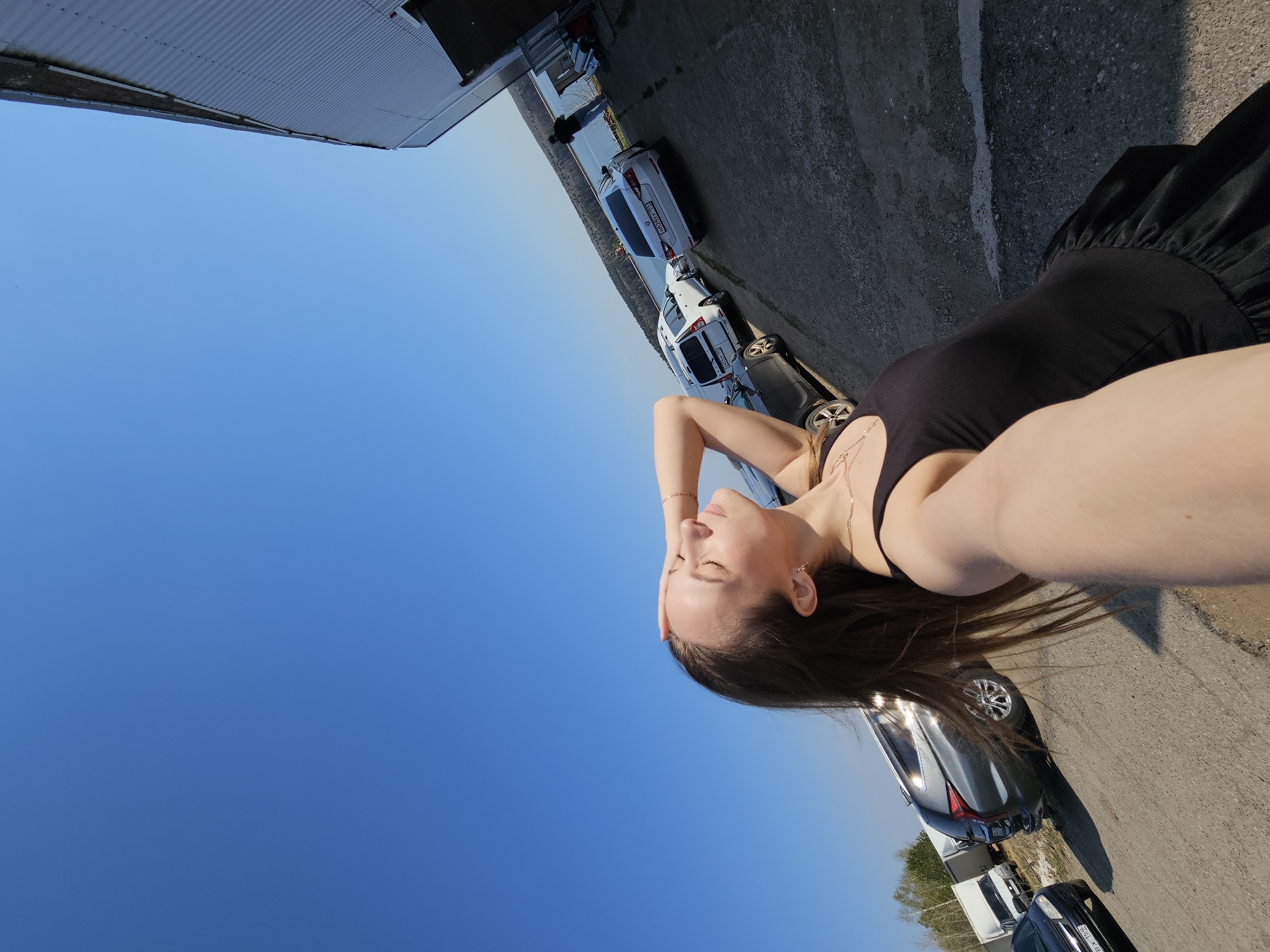 A woman in a black dress poses for a selfie outdoors. Cars are parked nearby. The image was likely taken in Chaikovsky, Russia.