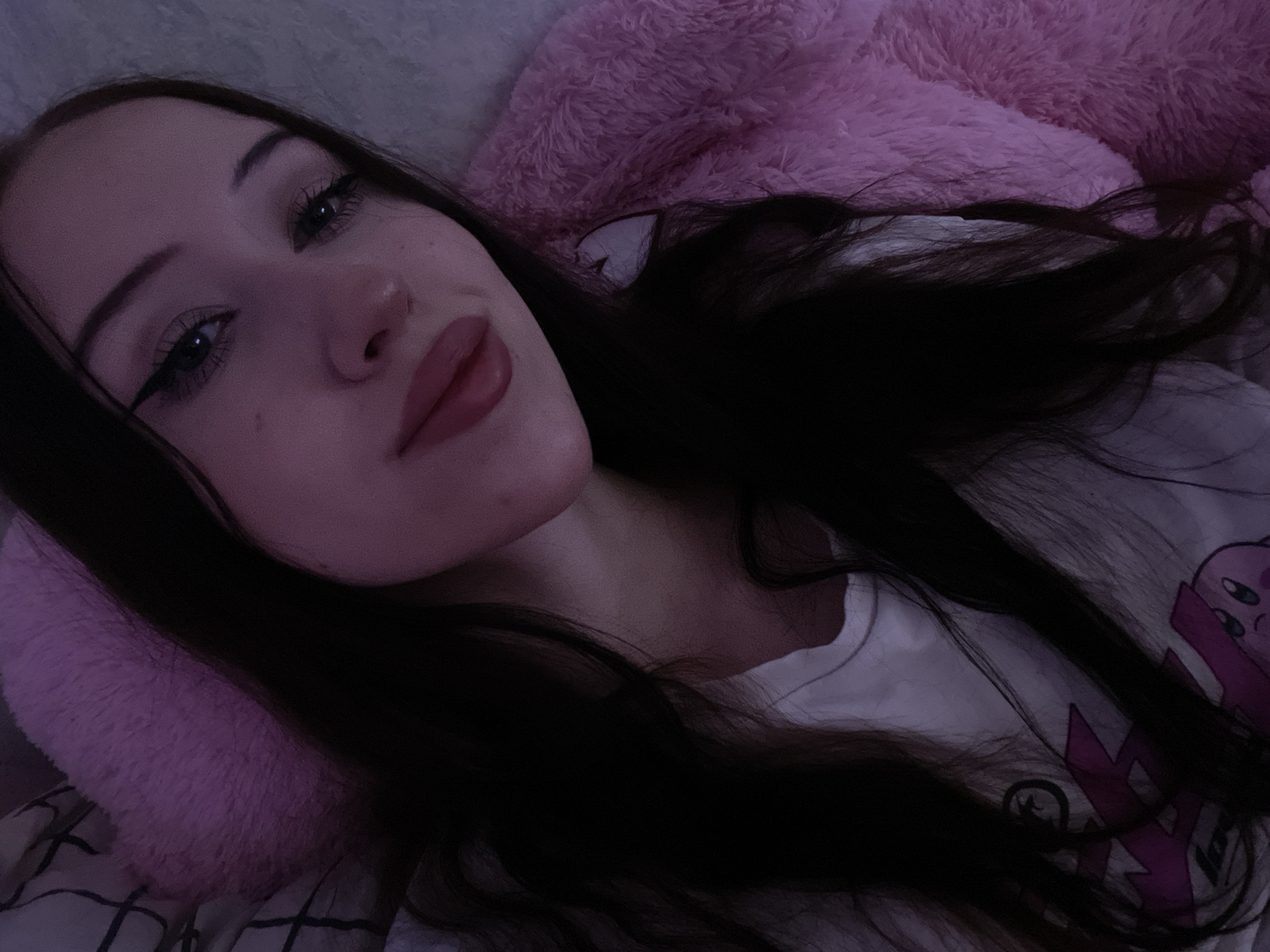 The image shows a close-up, low-angle selfie of a young woman with long, dark hair. She is wearing a white t-shirt with a graphic print. Her eyes are made up with eyeliner and mascara. She has full lips and fair skin. She is lying down, with a fluffy pink object, possibly a blanket or pillow, behind her head and to her right. The lighting is dim and casts a purple hue over the scene. The background appears to be a textured surface, perhaps a bed or sofa. There is no indication of the location in Votkinsk, Russia.
