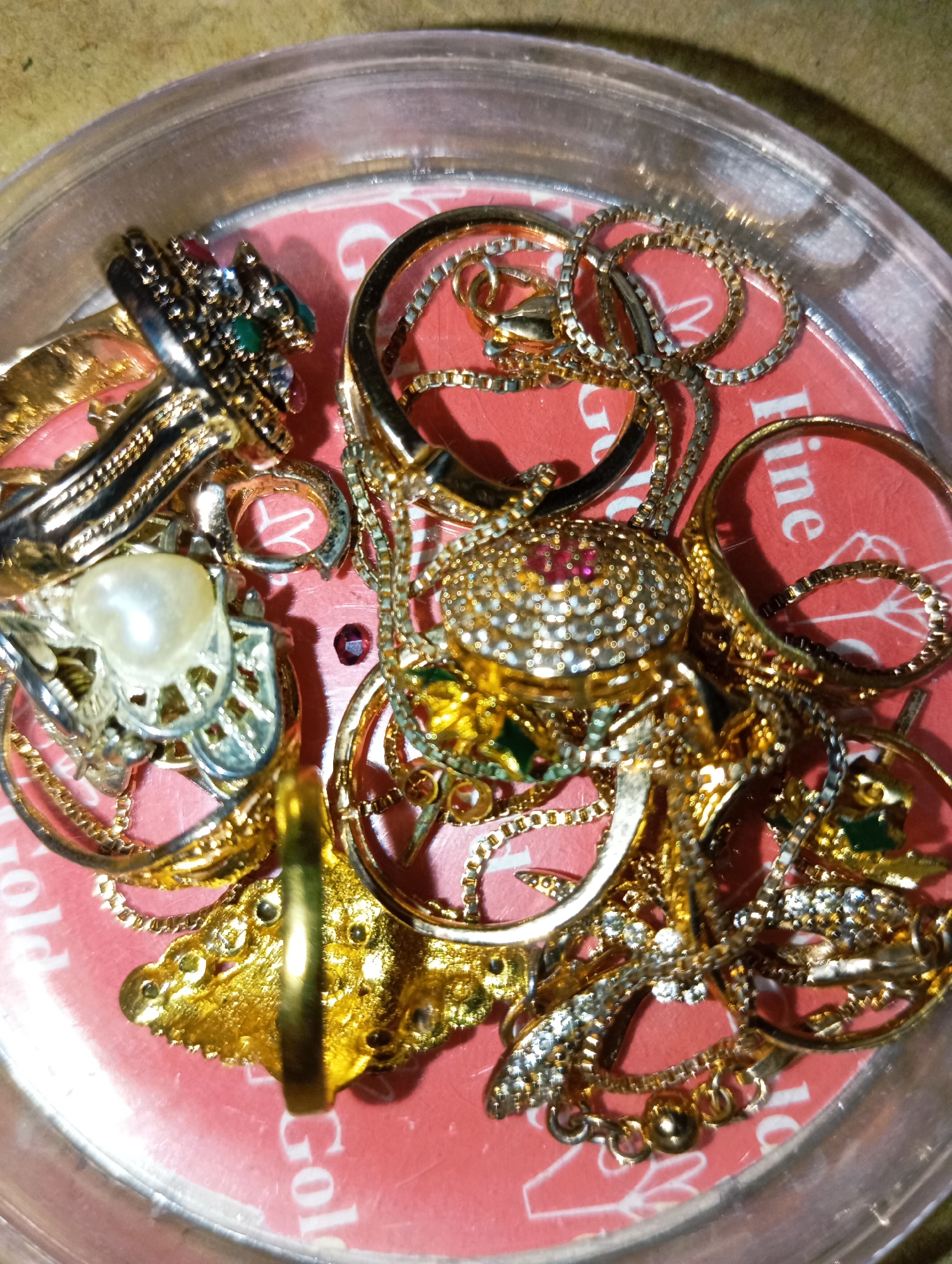 The image shows a close-up, overhead view of a collection of gold and silver-toned jewelry piled inside a clear plastic container. The jewelry includes numerous rings, some with elaborate designs and gemstones in red, green, and white. There are also several delicate gold chains and a pearl pendant. The items are jumbled together on a red surface with white text. Part of the text "Gold" is visible in the lower left corner, and "Fine" is visible in the upper right. The lighting suggests an indoor setting, with reflections on the metallic surfaces of the jewelry. The overall impression is one of abundance and variety within the collection.