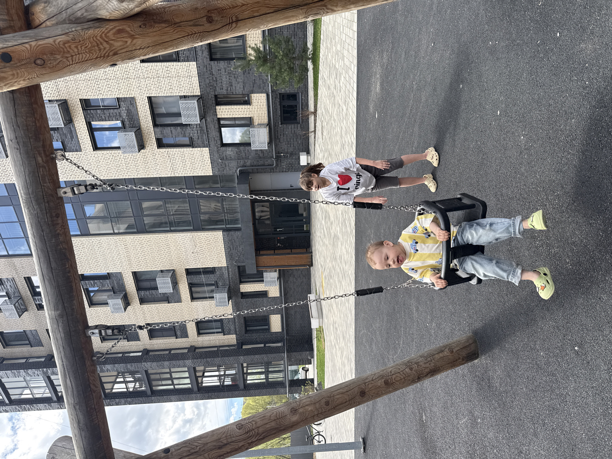 The image depicts two children on a playground, likely in the city of Rusanovo, Russia. One child is on a swing, wearing a yellow striped shirt, jeans, and lime-green Crocs. The other child, presumably pushing the swing, is wearing a white shirt with a heart graphic and gray shorts. The playground is located near a modern apartment building with a facade featuring a mix of light beige and dark gray brick. A wooden structure, possibly part of the swing set, is visible in the foreground. The ground is paved with asphalt.