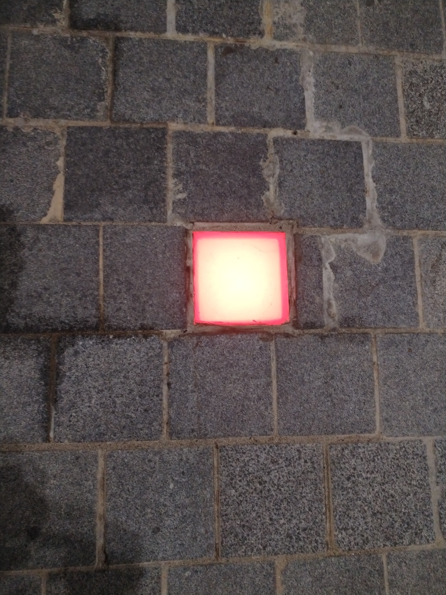 The image shows a section of a grey brick wall with a square, illuminated light fixture embedded in it. The fixture has a bright white center and a pink border. The brickwork is roughly textured, and the mortar between the bricks is visible. The scene appears to be located in an urban environment, potentially in Nha Trang Ward, Vietnam, though this cannot be verified from the image alone. There are no people visible.