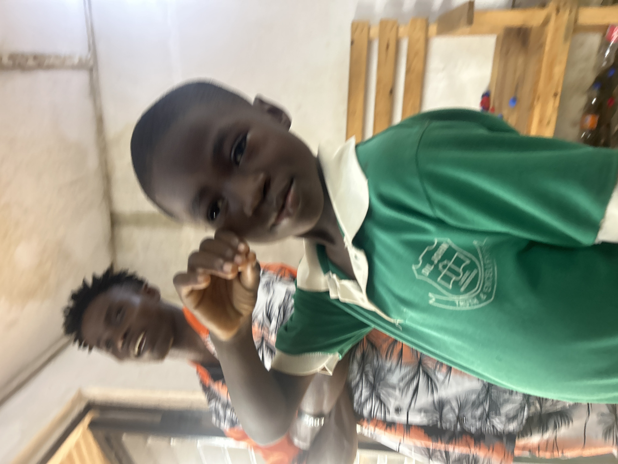 The image shows two young Black boys indoors. The boy in the foreground is looking towards the camera and has his right hand near his face. He is wearing a green polo shirt with a white collar and a crest on the left side of his chest. The crest is white and appears to be a shield with some symbols inside. The other boy, in the background and slightly out of focus, is smiling and looking towards his left. He is wearing a patterned shirt with black, white, and orange colors.
The setting appears to be a room with light-colored walls and wooden structures in the background, possibly shelves or a partition made of wooden planks. There is also a hint of a Christmas tree with decorations on the far right, suggesting the photo may have been taken around the holiday season. The lighting is somewhat dim, creating shadows. The overall mood seems casual and candid. No time of day or weather can be determined. No discernible text is visible apart from the crest on the shirt.