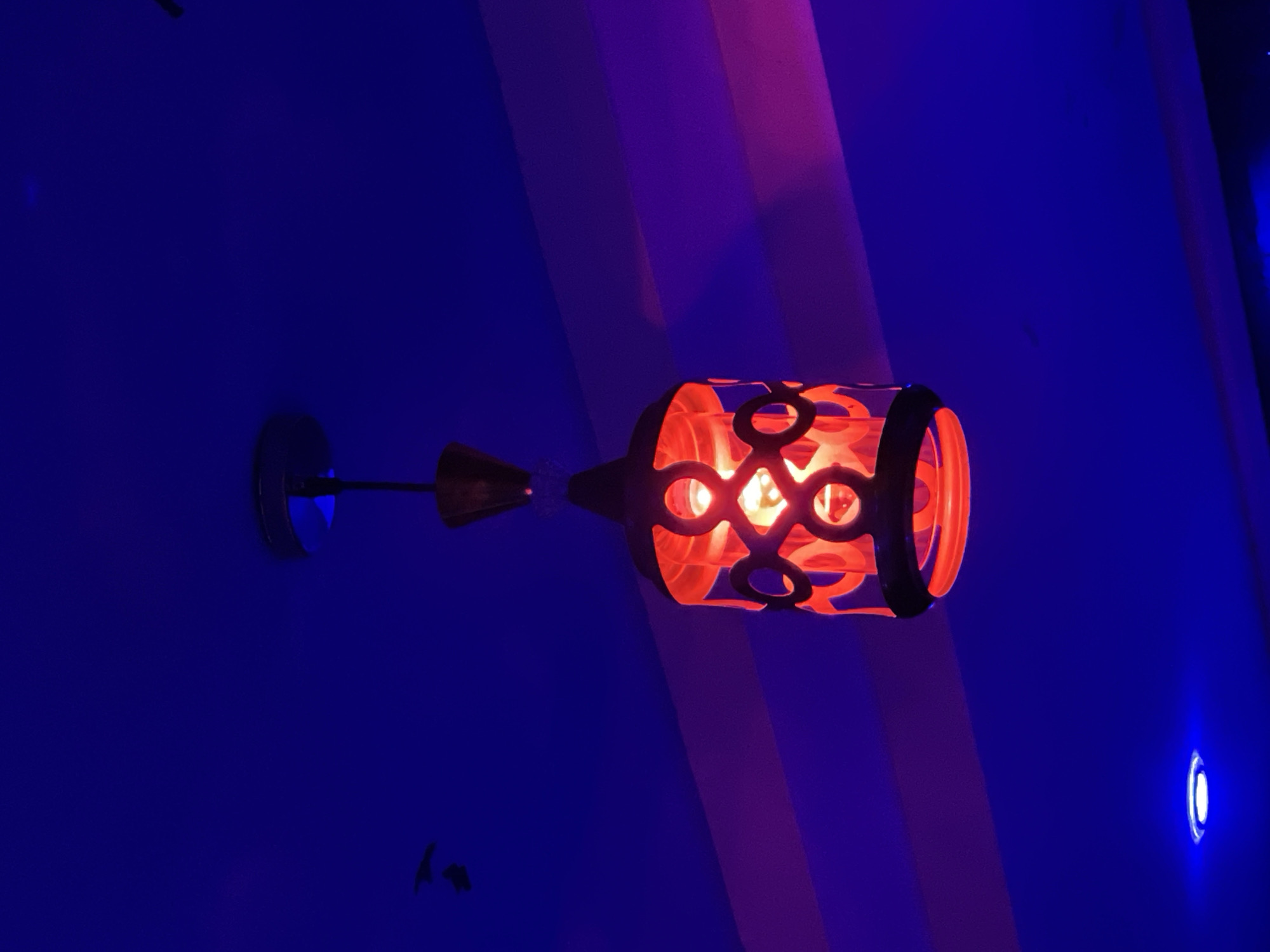The image shows a decorative lamp with an orange, patterned shade, illuminated by a warm light from within. The lamp is mounted on a dark surface, likely a wall, which is painted in a deep blue color, with a section of the wall showing a gradient of purple and pink hues, possibly due to ambient lighting or a painted architectural feature. The lamp itself has an intricate design with circular cutouts. A thin, dark arm extends from the lamp to a circular mounting base.
In the background, the blue wall continues, with subtle variations in shade suggesting contours or architectural elements. To the right of the lamp, a small, bright blue light is visible, possibly another light fixture or a reflection. The overall mood is atmospheric and perhaps a bit dramatic, with the interplay of the warm orange light from the lamp and the cool blue and purple tones of the wall. There is no visible text, people, or specific indication of the time of day or weather conditions, beyond the implication of an indoor setting with controlled lighting. The scene appears to be in a place with architectural detail that creates varied lighting effects.