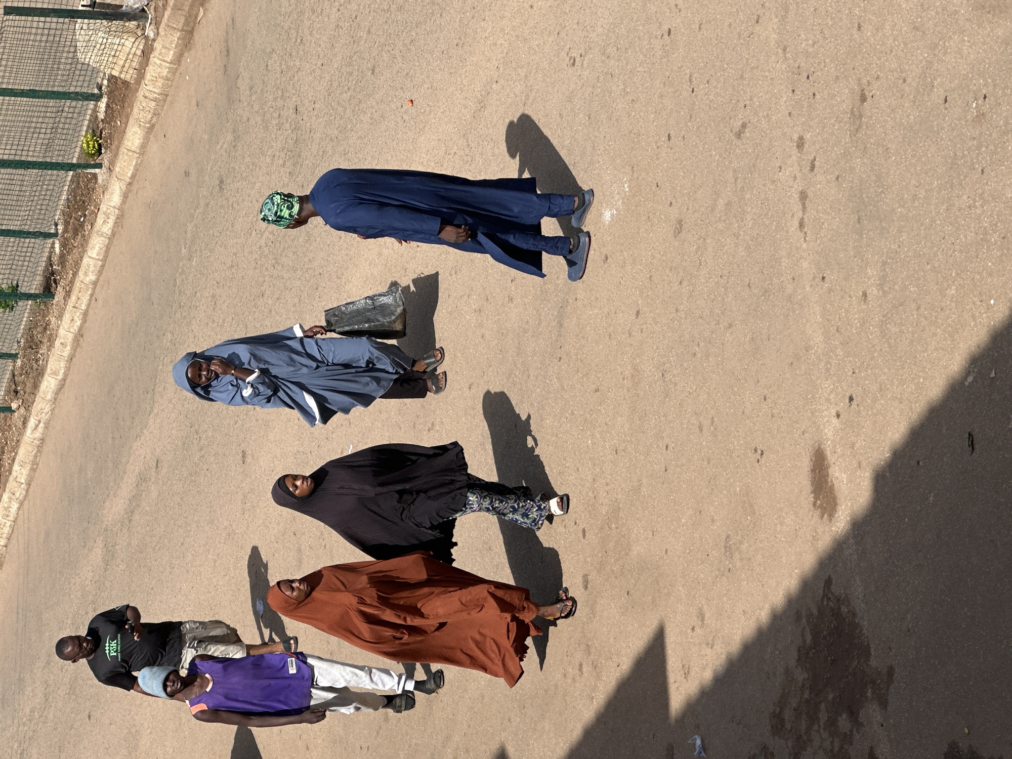 In Bukuru, Nigeria, on a paved street, a group of individuals walks outdoors. A man wearing a black t-shirt with white lettering and glasses walks toward the left, holding a dark smartphone in his right hand. Next to him, a young person in a purple tank top and a light blue beanie walks forward, holding their arms at their sides. Following them, a woman in a burnt orange headscarf and dress walks with her head down. To her right, another woman wears a black headscarf and patterned trousers, and she looks to her left. Further to the right, a person in a dark blue kaftan and a green and black patterned hat walks away from the viewer, carrying a black plastic bag in their right hand. The ground is a light brown asphalt surface with scattered dark spots. A green metal fence lines the top of the image, with dry vegetation and a concrete curb beneath it. Shadows are cast across the ground, indicating a bright, sunny day.