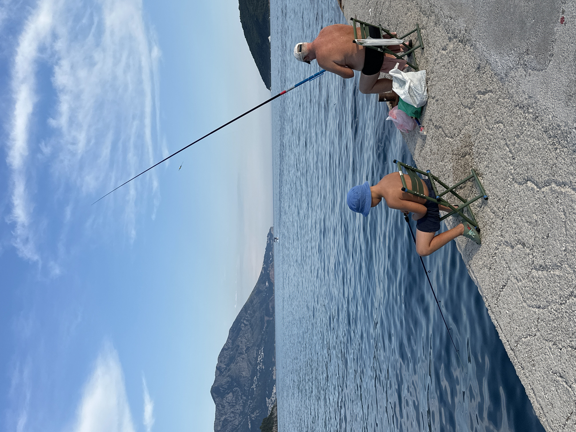 Two individuals, an adult male and a boy, are fishing from a concrete pier in Budva, Montenegro. The adult male, seated on a green folding chair, wears black swim trunks and a white baseball cap. The boy, also seated on a similar chair, wears a blue bucket hat and dark shorts. Both are holding fishing rods and appear to be focused on the water.
The sea is a deep blue, with gentle ripples on the surface. In the background, a large, mountainous coastline stretches across the horizon, dotted with buildings. The sky is a clear blue, with scattered white clouds. Bags are placed on the pier near the fishermen, suggesting they have brought supplies with them. The scene captures a moment of leisure and outdoor activity by the Adriatic Sea.