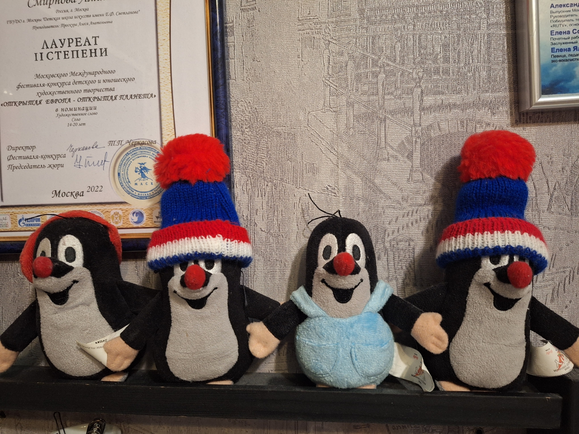 Four plush toys, resembling the character "The Little Mole" (Krtek), are displayed on a dark shelf against a textured wallpaper. Each mole has a black body, grey belly, and a prominent red nose.
The two moles on the left wear solid red knitted hats. The two moles on the right wear red, white, and blue striped knitted hats with red pom-poms. The third mole from the left is also dressed in light blue overalls. Tags are visible on some of the toys, with partially readable text including "MILCE", "1900", and "MORAVSKA BANO", likely indicating their origin from a Czech manufacturer.
Behind the moles, on the left, hangs a framed certificate written in Russian. The certificate states "Россия, г. Москва" (Russia, Moscow) and is from "ГБУДО г. Москвы 'Детская школа искусств имени Е.Ф. Светланова'" (Moscow Children's Art School named after E.F. Svetlanova). It designates a "ЛАУРЕАТ ІІ СТЕПЕНИ" (LAUREATE OF II DEGREE) from the "Московского Международного фестиваля-конкурса детского и юношеского художественного творчества" (Moscow International festival-competition of children's and youth artistic creativity) titled "«ОТКРЫТАЯ ЕВРОПА - ОТКРЫТАЯ ПЛАНЕТА»" (OPEN EUROPE - OPEN PLANET) in the nomination "Художественное слово Соло 14-20 лет" (Artistic word Solo 14-20 years old). It mentions "Преподаватель: Проскура Алеся Анатольевна" (Teacher: Proskura Alesya Anatolyevna) and is signed by the "Директор Фестиваля-конкурса Т.П. Черкасова" (Director of the Festival-competition T.P. Cherkasova) and "Председатель жюри" (Chairman of the Jury). The date "Москва 2022" is visible, along with a circular seal bearing "МАСК".
On the right, another framed document is partially visible, also containing text in Russian. This document appears to list individuals and their achievements or roles, including "Александр", "Елена Со", and "Елена Я". Keywords like "Выпускник Мос" (Graduate of Mos...), "Руководитель" (Head), "Победитель те" (Winner of the...), "RUTV", "Почетный рабе" (Honorable worker), "Заслуженный" (Honored), "Певица, педаг" (Singer, pedagogue), and "эко-вокалист" (eco-vocalist) are discernible. The background features a light-colored wallpaper with an embossed pattern.