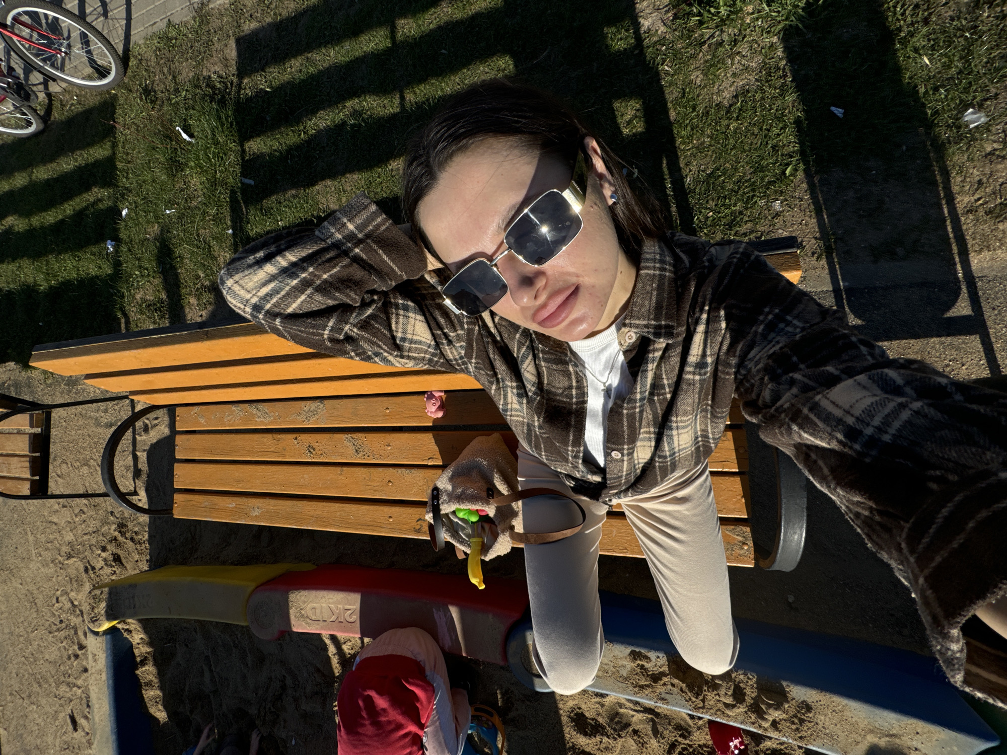 A high-angle, selfie-style shot captures a woman with brown hair wearing sunglasses and a plaid shirt. She is seated on a wooden bench located in what appears to be a playground or park area. Part of a playground structure, with elements colored red, yellow, and blue, is visible beneath the bench. A bicycle is partially visible in the upper left corner of the frame. The background consists of grass and shadows, suggesting a sunny day. The location may be in the city of Odintsovo, Russia.